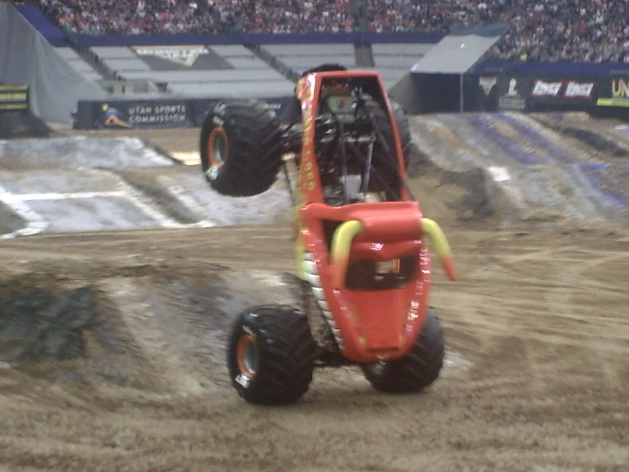 An orange monster truck with large tires is in mid-air at a stadium dirt arena during a monster truck event, with a crowd of spectators in the background.