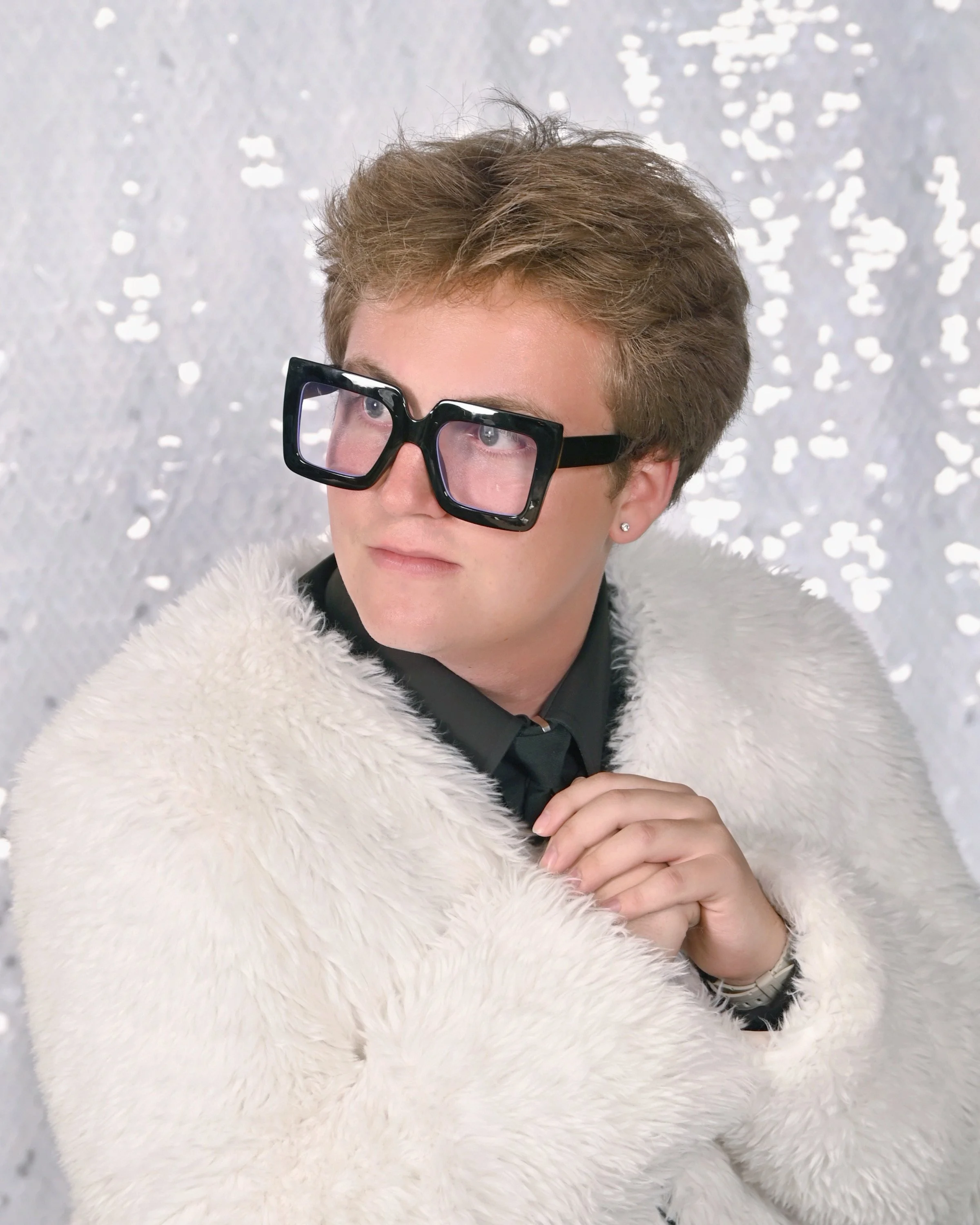 Person with short brown hair wearing black glasses, a black shirt with a collar, a white fluffy coat, and a silver watch, standing in front of a sparkly white textured background.