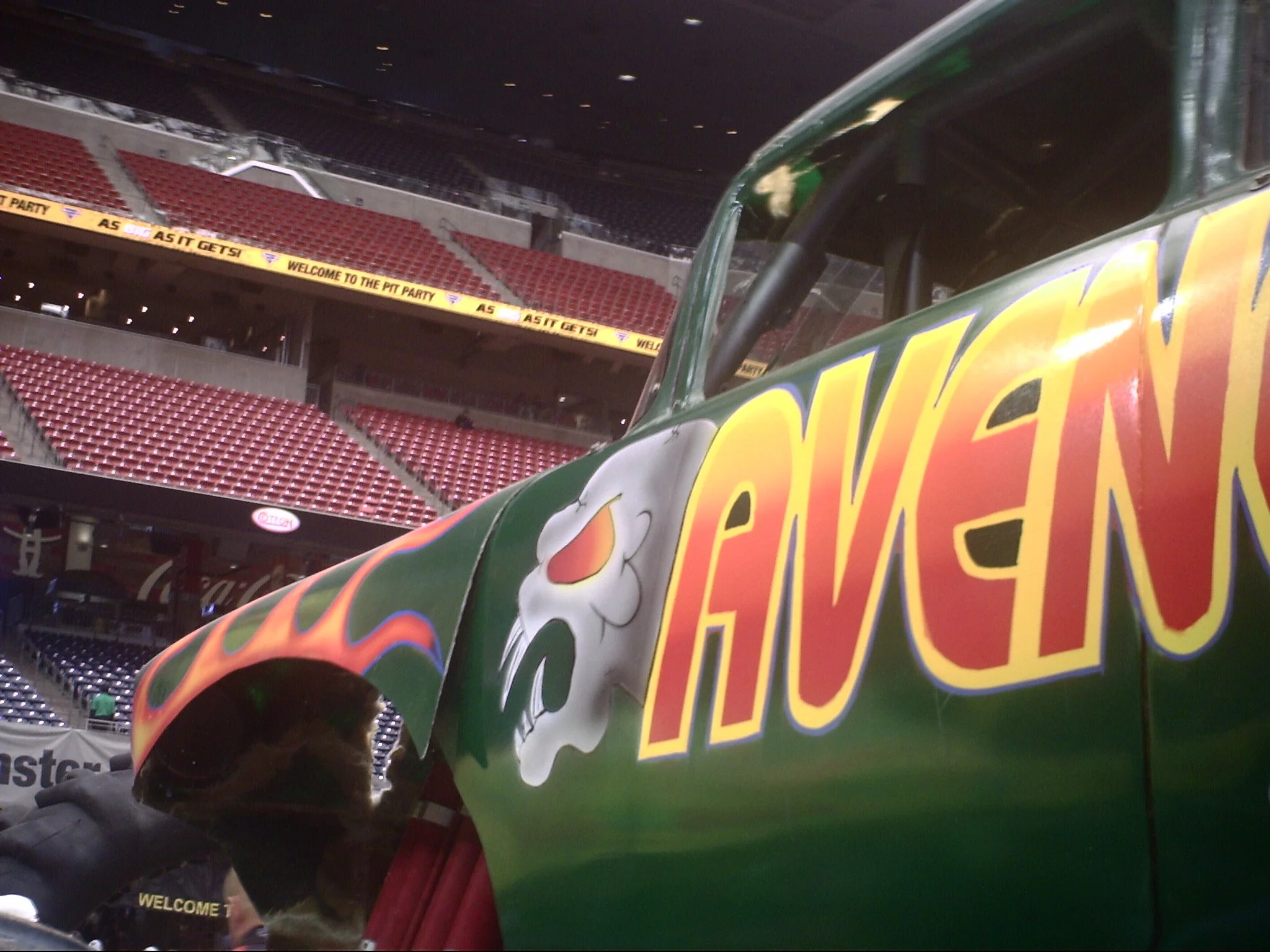 Part of a colorful race car or promotional vehicle inside a stadium, with red stadium seats and a yellow banner that reads "WELCOME TO THE PIT PARTY." A person is visible in the lower left corner of the image.