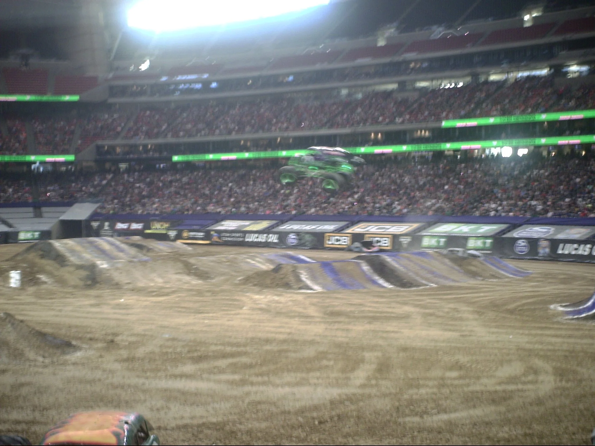 View of a dirt motocross track with jumps and a large stadium filled with spectators in the background. A vehicle is airborne above one of the jumps.