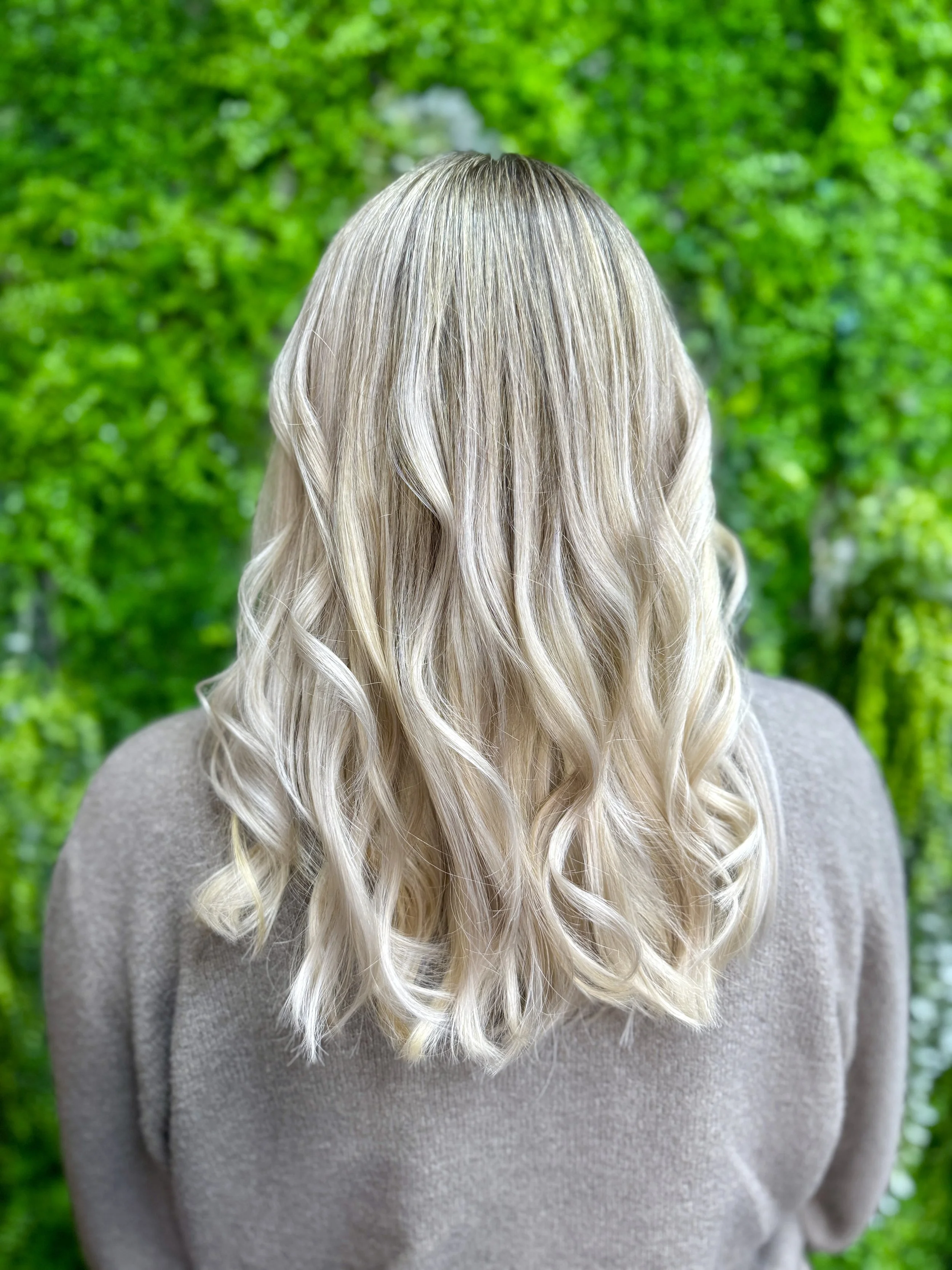 Der Rücken einer Person mit langen, glatten, blonden Haaren, die einen hellgrauen Pullover trägt, vor einem grünen, bewaldeten Hintergrund.