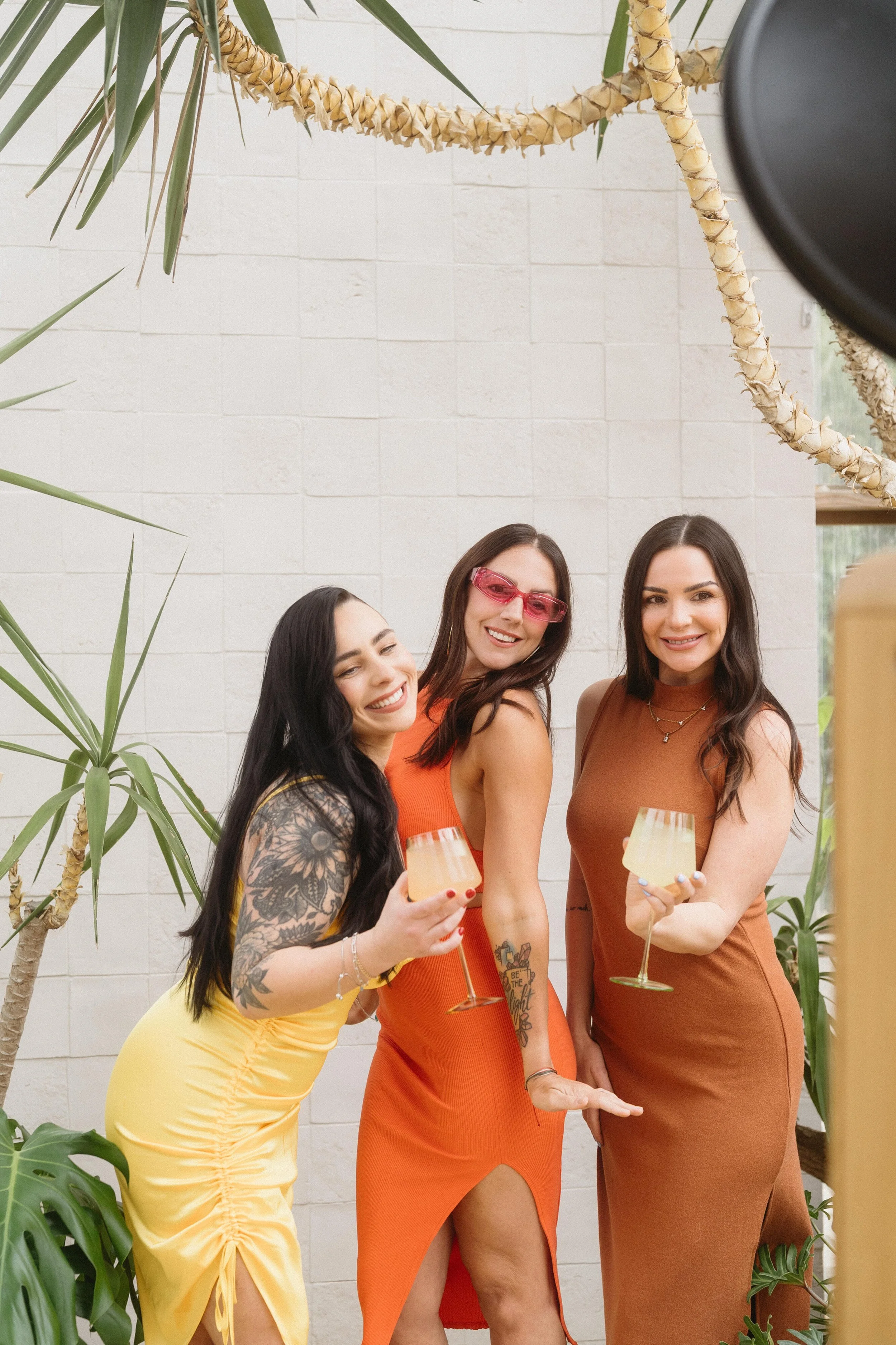 Three women smiling and holding drinks at a party, standing in front of plants and a white wall.