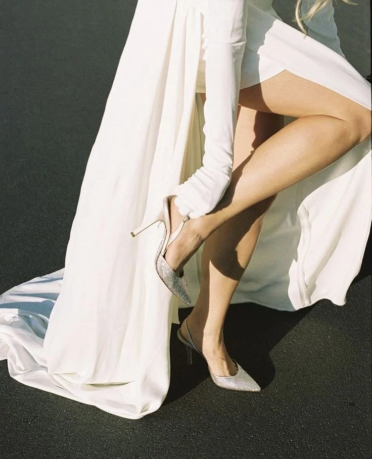 Close-up of a woman in a white dress with silver high heels, lifting her leg to reveal her shoe and part of her thigh. She is seated on a white fabric draped over a black surface.