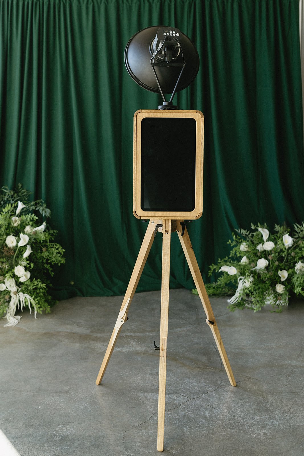 A vintage speaker on a tripod stand, with a large black satellite speaker and a smaller control unit on top, set against a green curtain backdrop with floral arrangements on the sides.