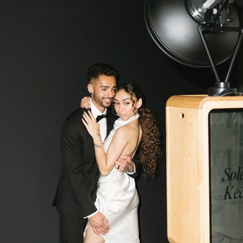 A man and woman in wedding attire posing together against a dark background, with a large film camera or spotlight partially visible to the right.