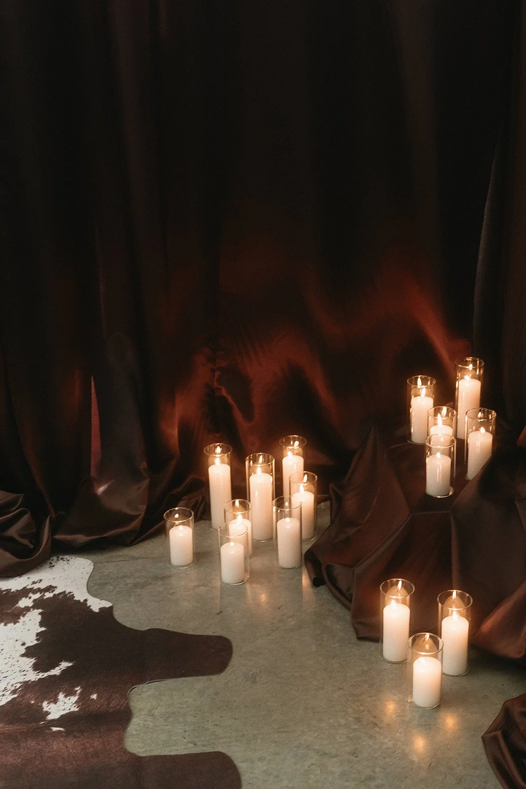Group of white pillar candles burning on a dark fabric backdrop, with a cowhide rug on the floor.