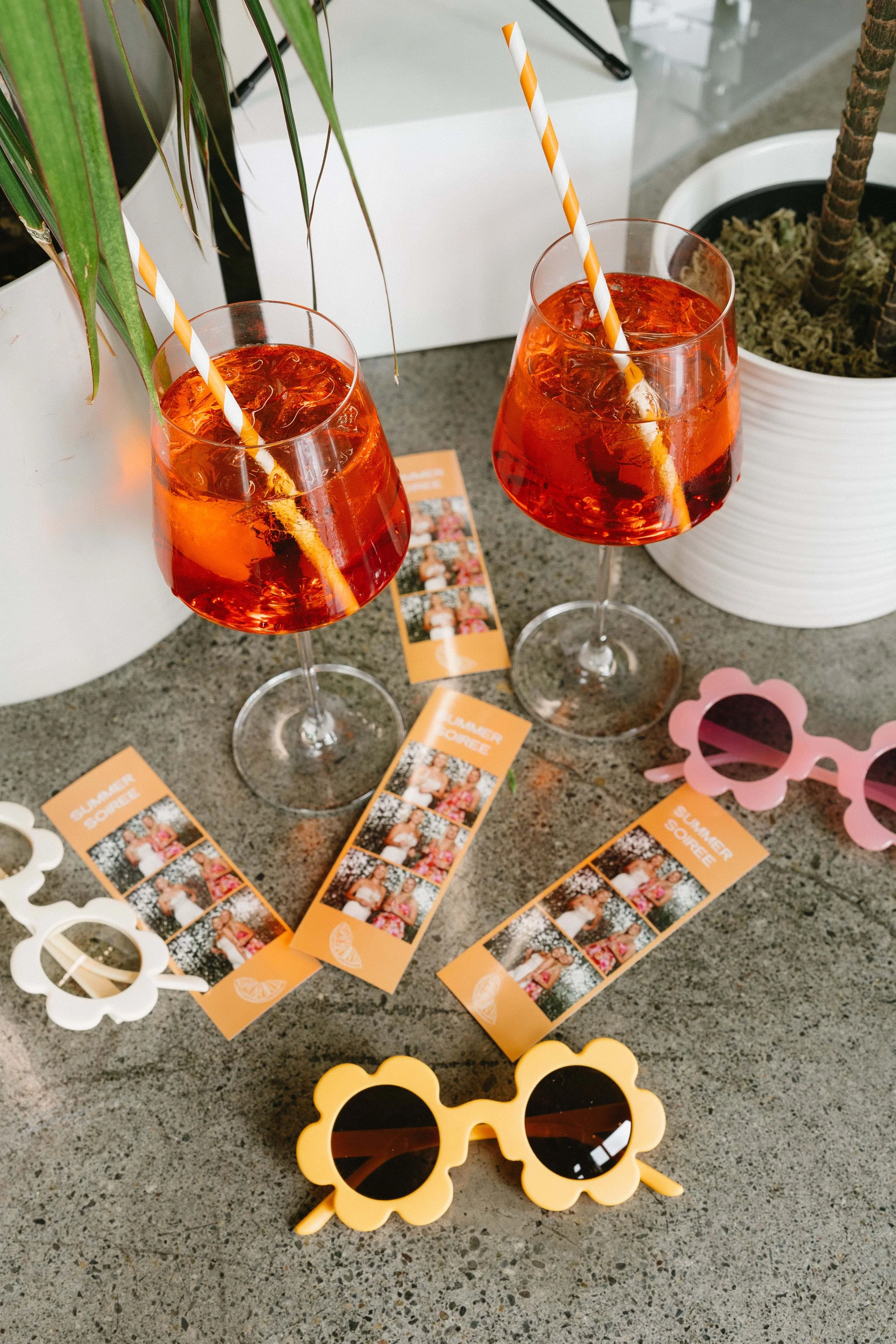 Two glasses of an orange-colored beverage with ice and striped straws on a table with party favors, including paper sunglasses and colorful cards, near potted plants.