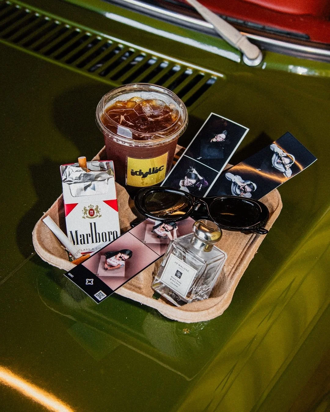 A tray with an iced coffee, a pack of Marlboro cigarettes, sunglasses, a perfume bottle, and several printed photographs, all on a green table.