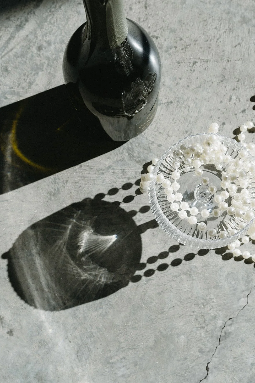 A black bottle with a green cleaning cloth on top, casting a shadow on a concrete surface beside a clear glass dish filled with white pearl-like beads.