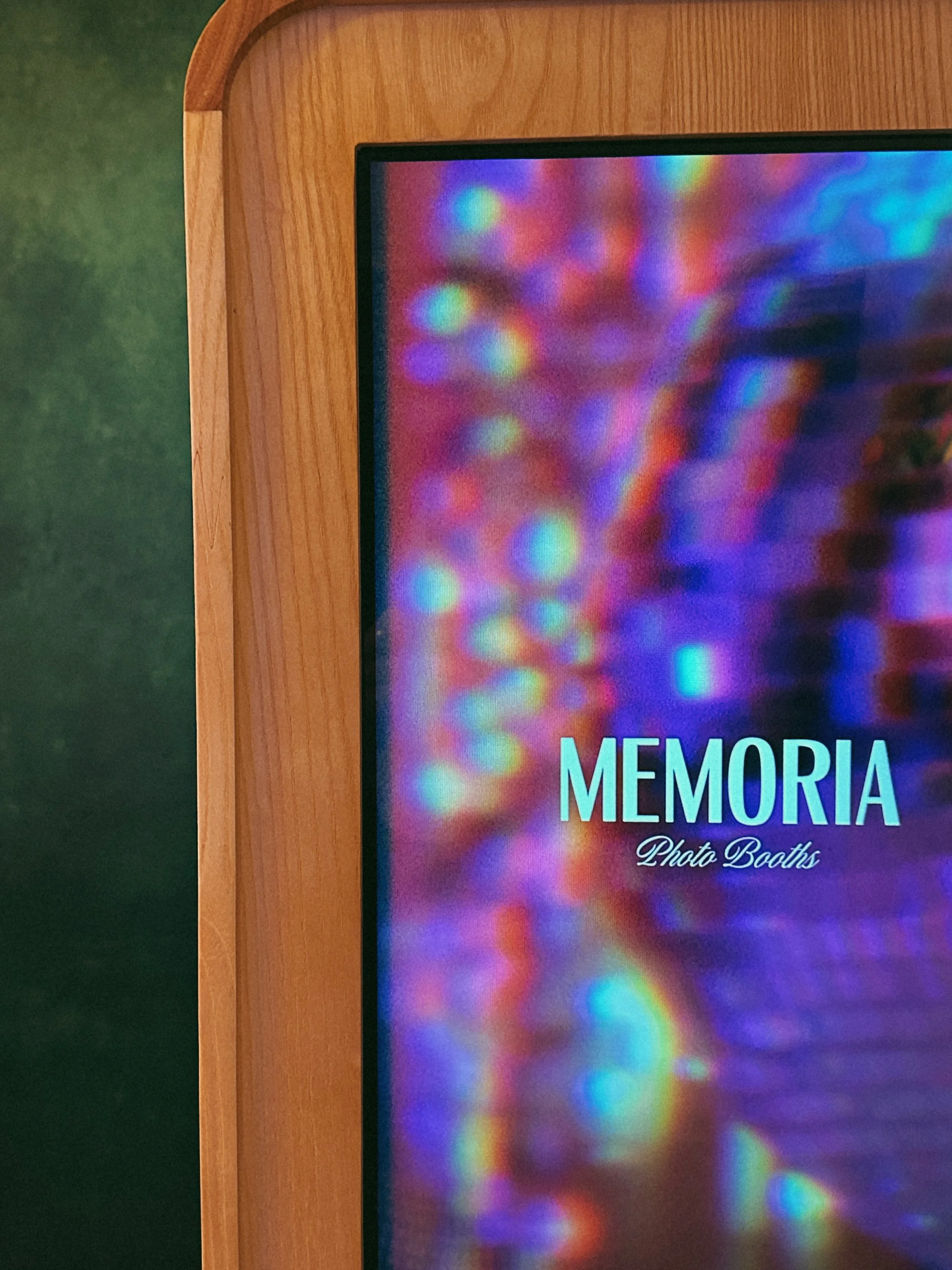 A digital photo booth screen framed in light wood, displaying colorful rainbow lights and the words "MEMORIA Photo Booths."