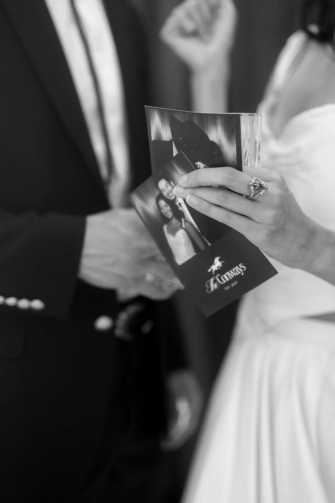 A person holding a photograph at a wedding, showing a woman in a wedding dress, with another person in a dark suit and a bride in a white wedding dress nearby.
