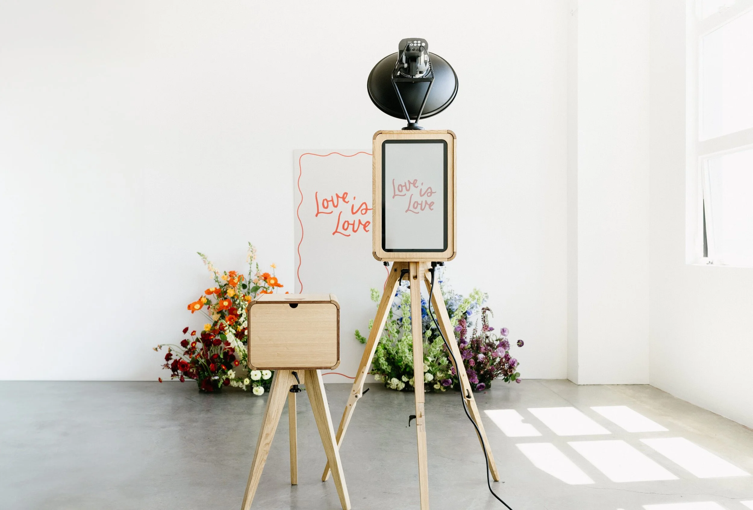 A photo of a photographer's setup with a camera on a tripod facing a white wall, surrounded by colorful flowers in a studio with natural light.