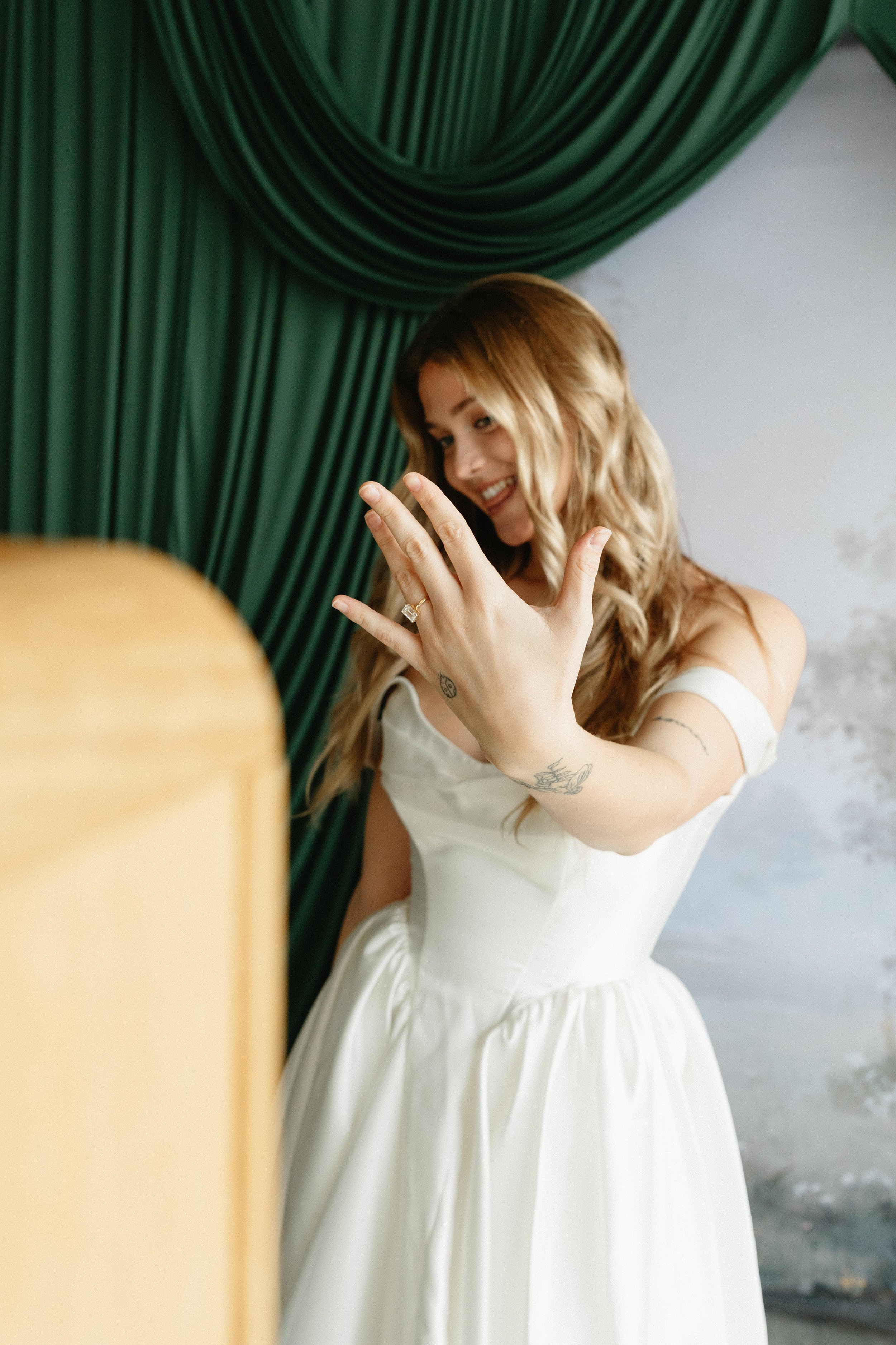 A woman with long, wavy hair smiling and looking at her hand, which has an engagement ring, in front of a green curtain and a white wall.