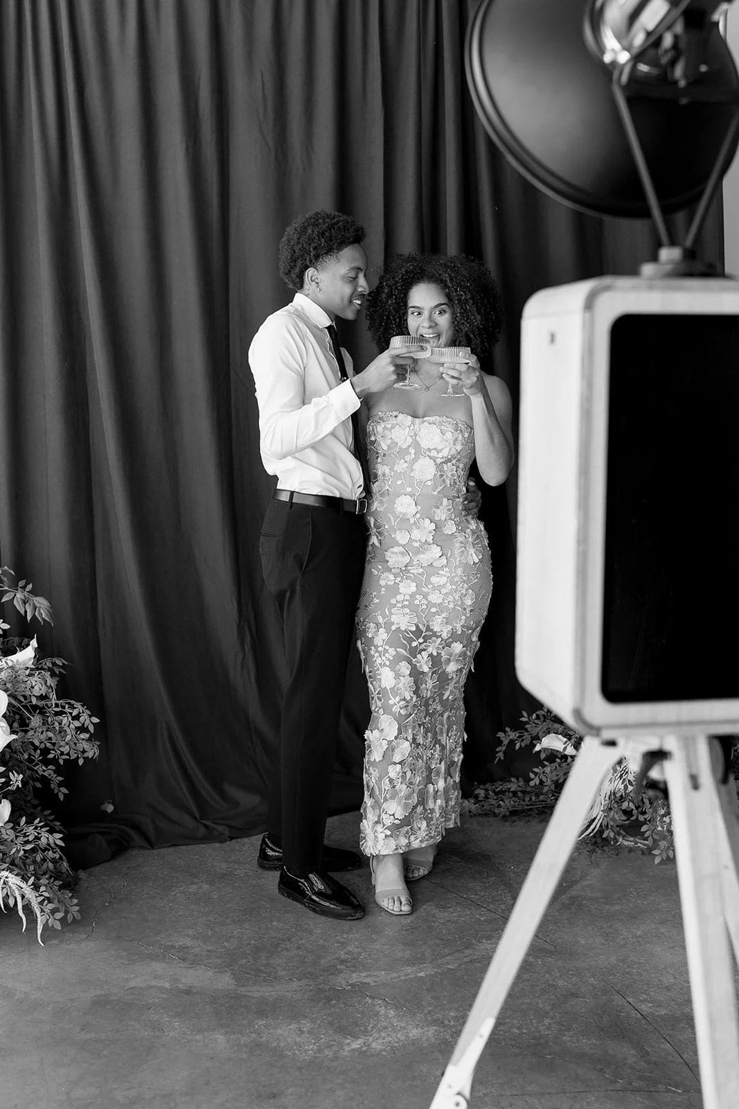 Two women, one in a floral dress and the other in a white shirt and tie, stand close together holding drinks and smiling for a photo while a camera records them. A curtain backdrop and some plants are visible.