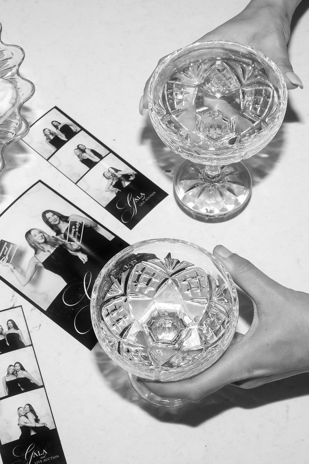 Two hands holding crystal wine glasses filled with a clear beverage, placed on a white surface with a photo booth strip and promotional flyers for a live auction event nearby.