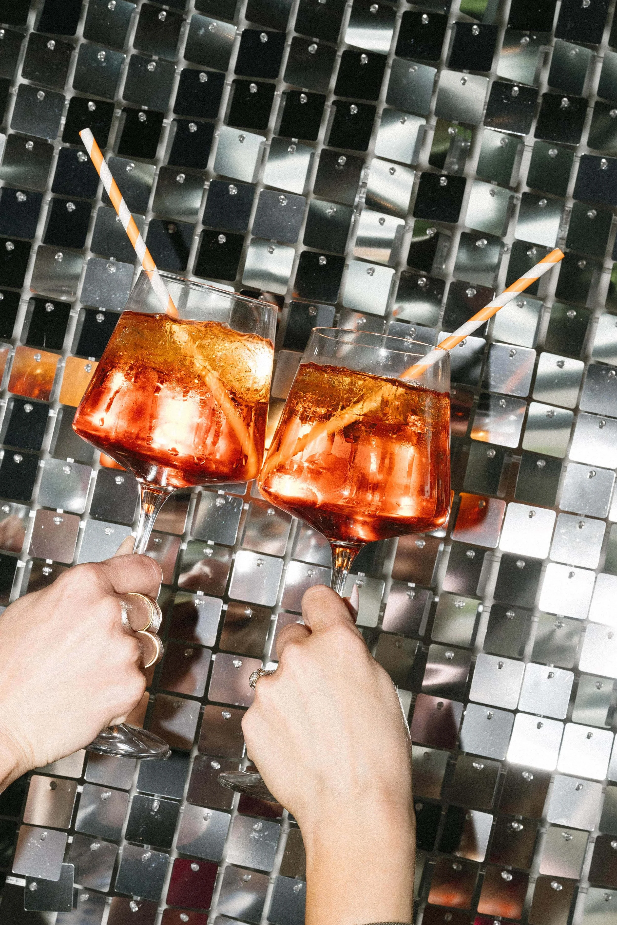 Two glasses of pink cocktail with ice, each with an orange and white striped straw, being held by two women with rings, against a shiny, mirrored, mosaic tile background.