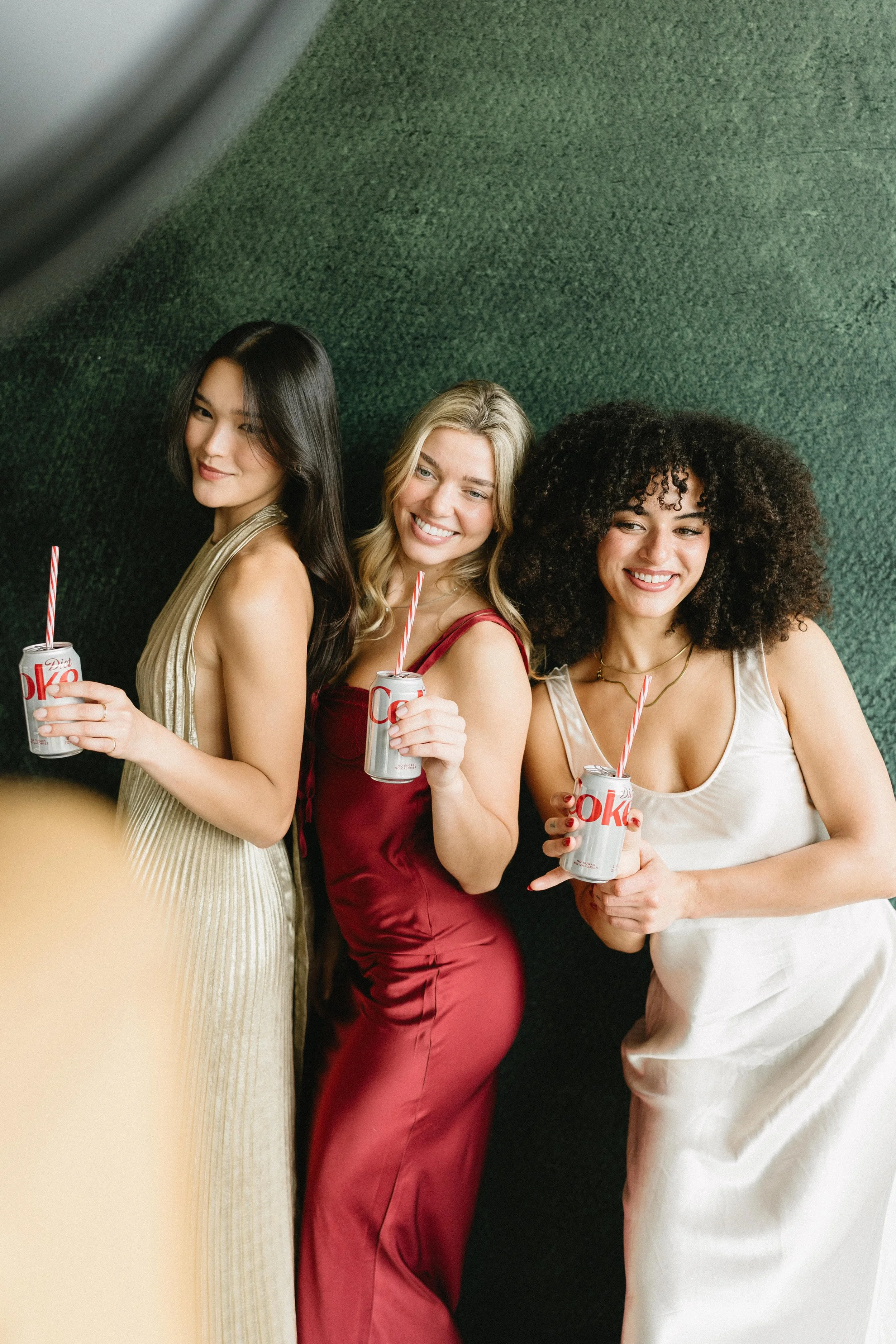 Three women standing together, smiling, holding cans of Diet Coke with straws, dressed in elegant dresses, against a textured green background.