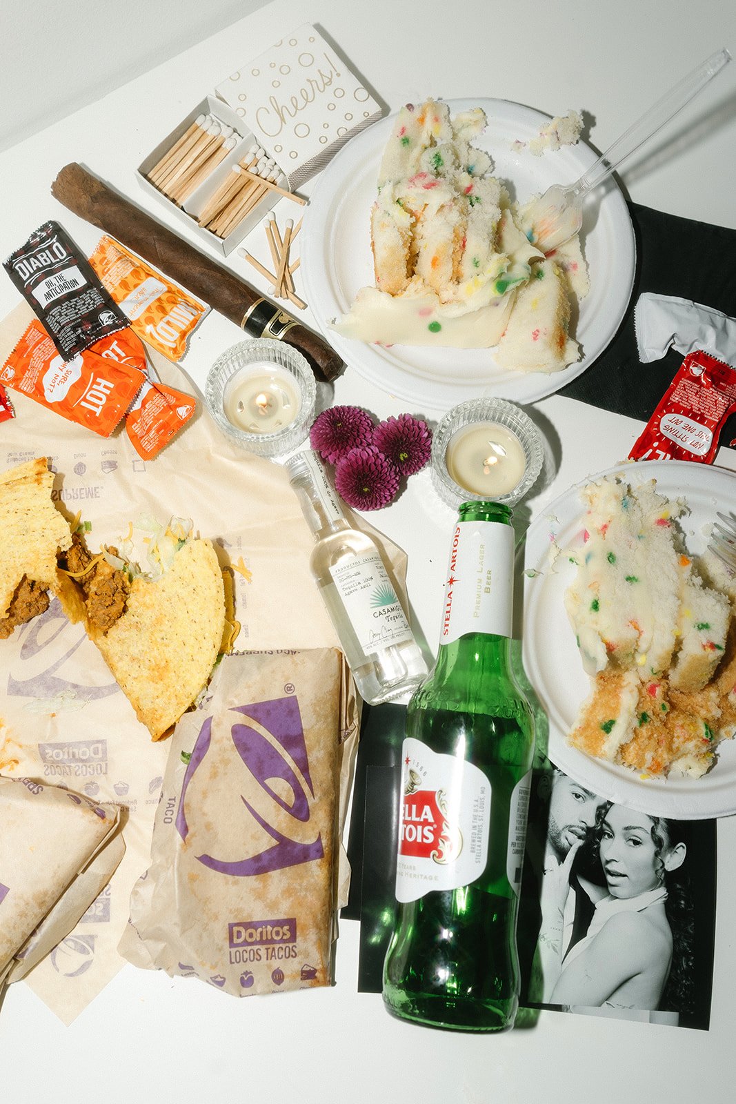 Table with cupcakes, candles, alcohol bottles, tacos, Doritos tacos, flowers, candles, and a black and white photo of a woman and man, suggesting a celebration or party.