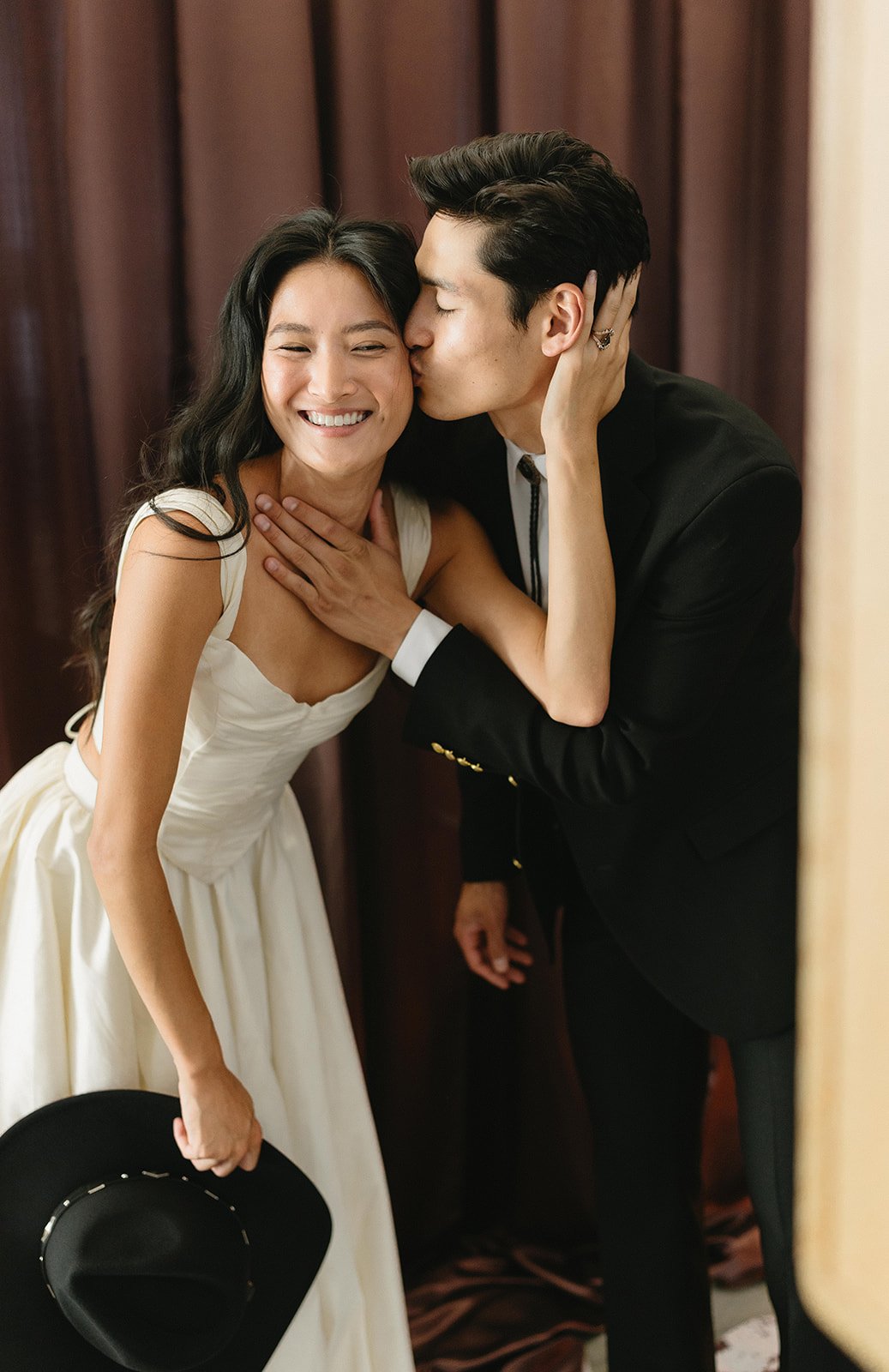 A couple sharing a kiss, with the woman smiling and the man kissing her on the cheek, inside a room with dark curtains.