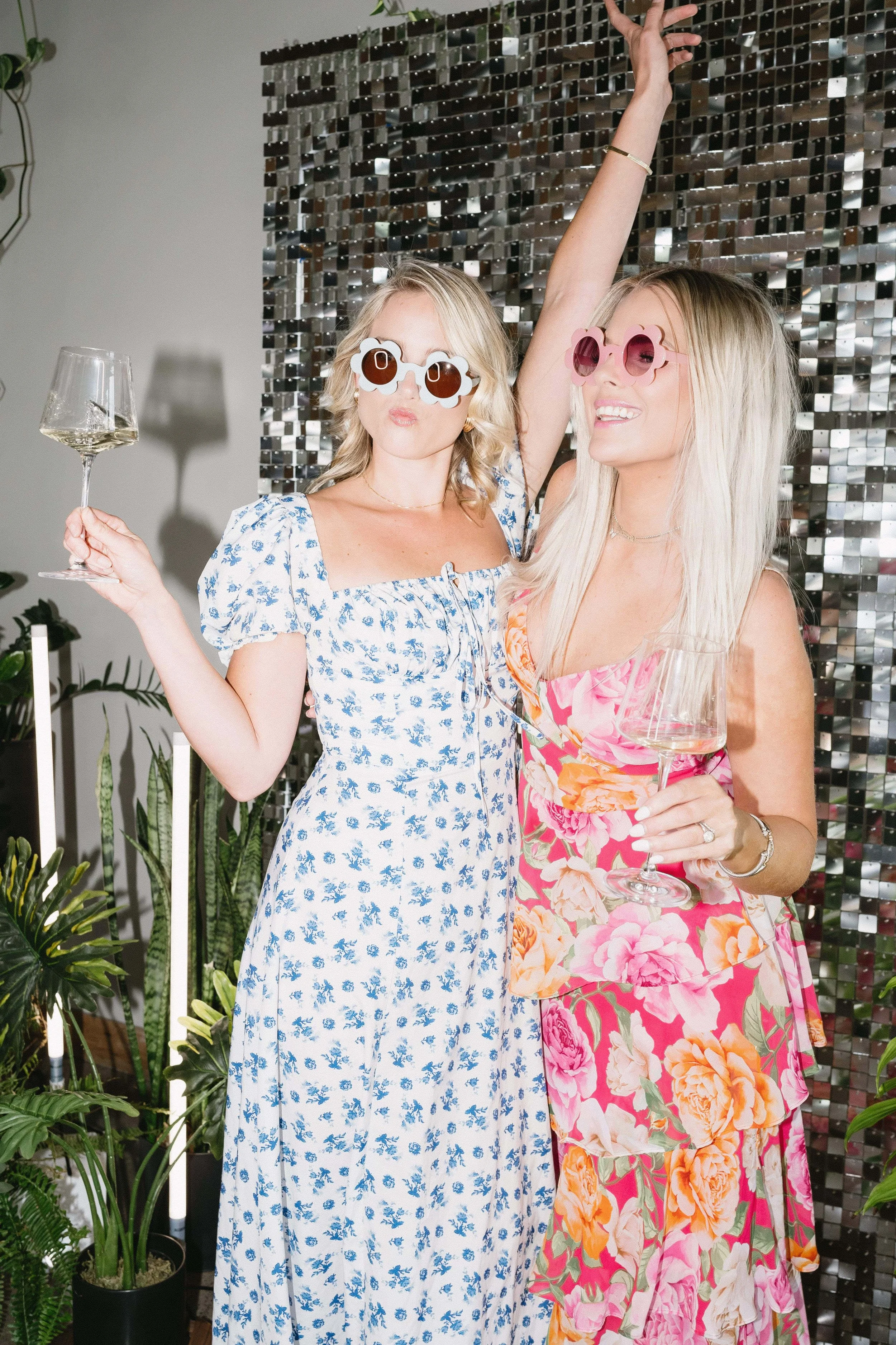Two women celebrating with wine at a party, wearing colorful floral dresses and stylish sunglasses, standing in front of a shiny, reflective backdrop, surrounded by green plants.