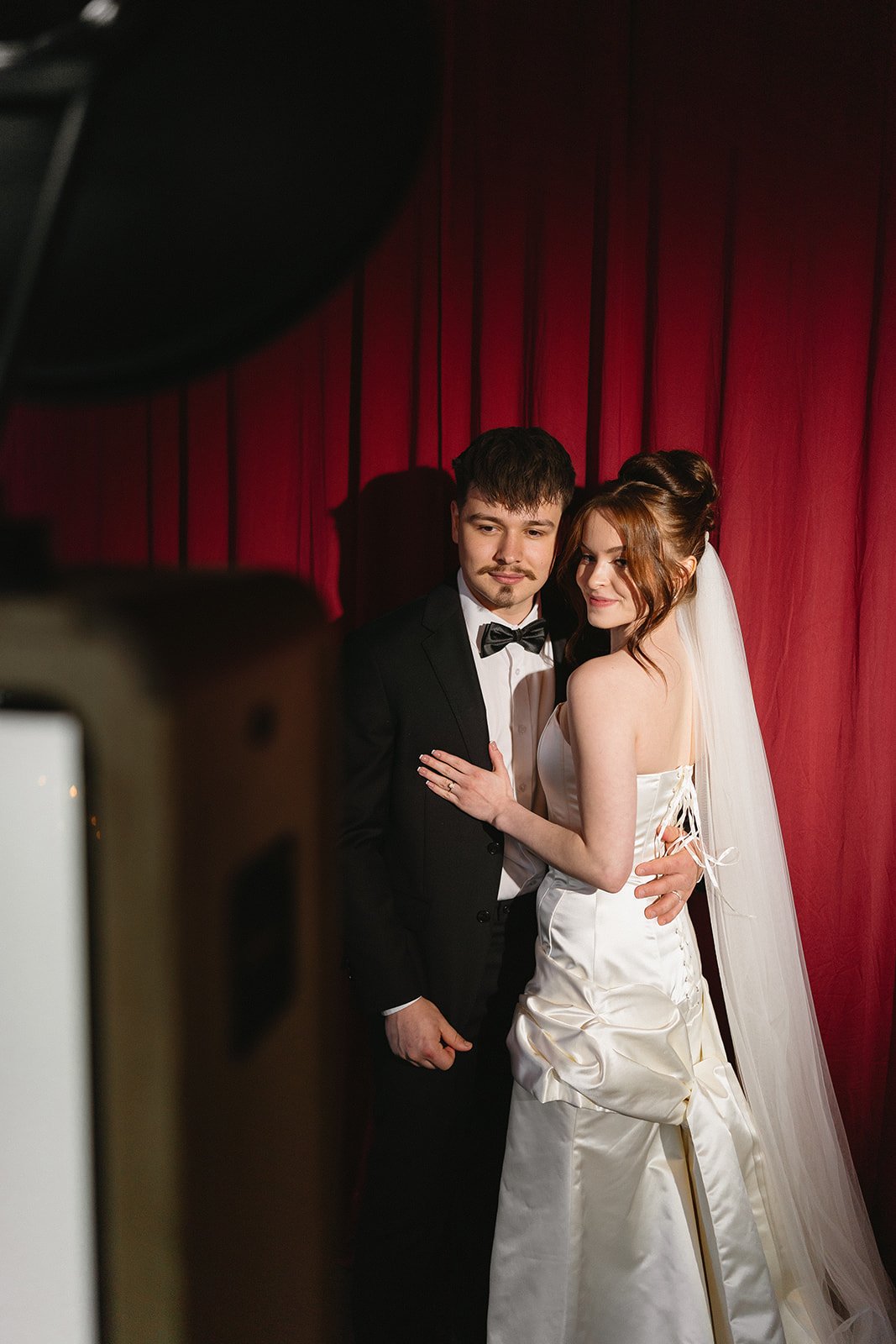 A bride and groom in wedding attire posing for a photo against a red curtain backdrop, with the bride wearing a strapless white wedding gown and veil, and the groom in a black tuxedo and bow tie, smiling gently.