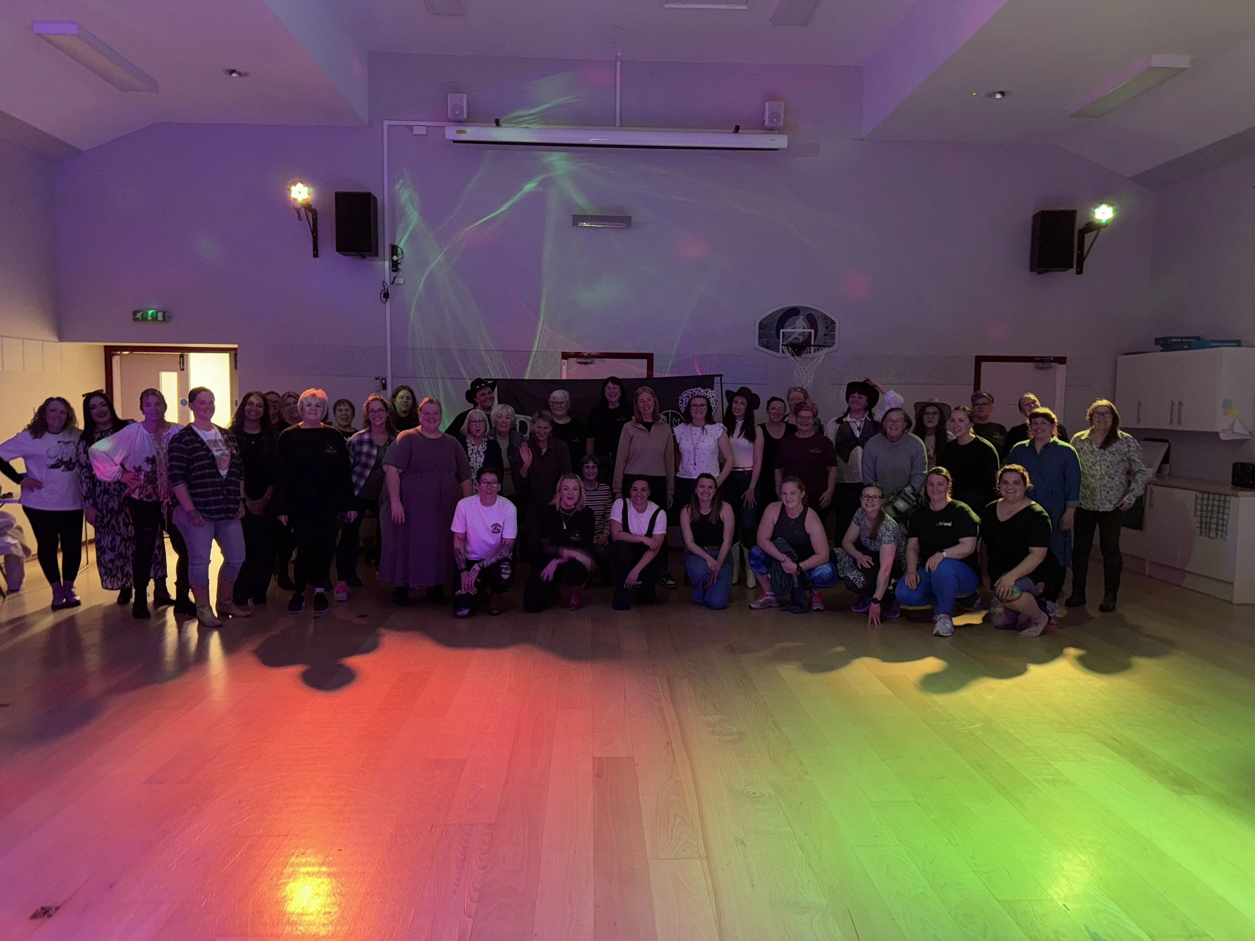 A large group of people, both men and women, standing together in a room with colorful lighting on the floor, possibly for a celebration or event.