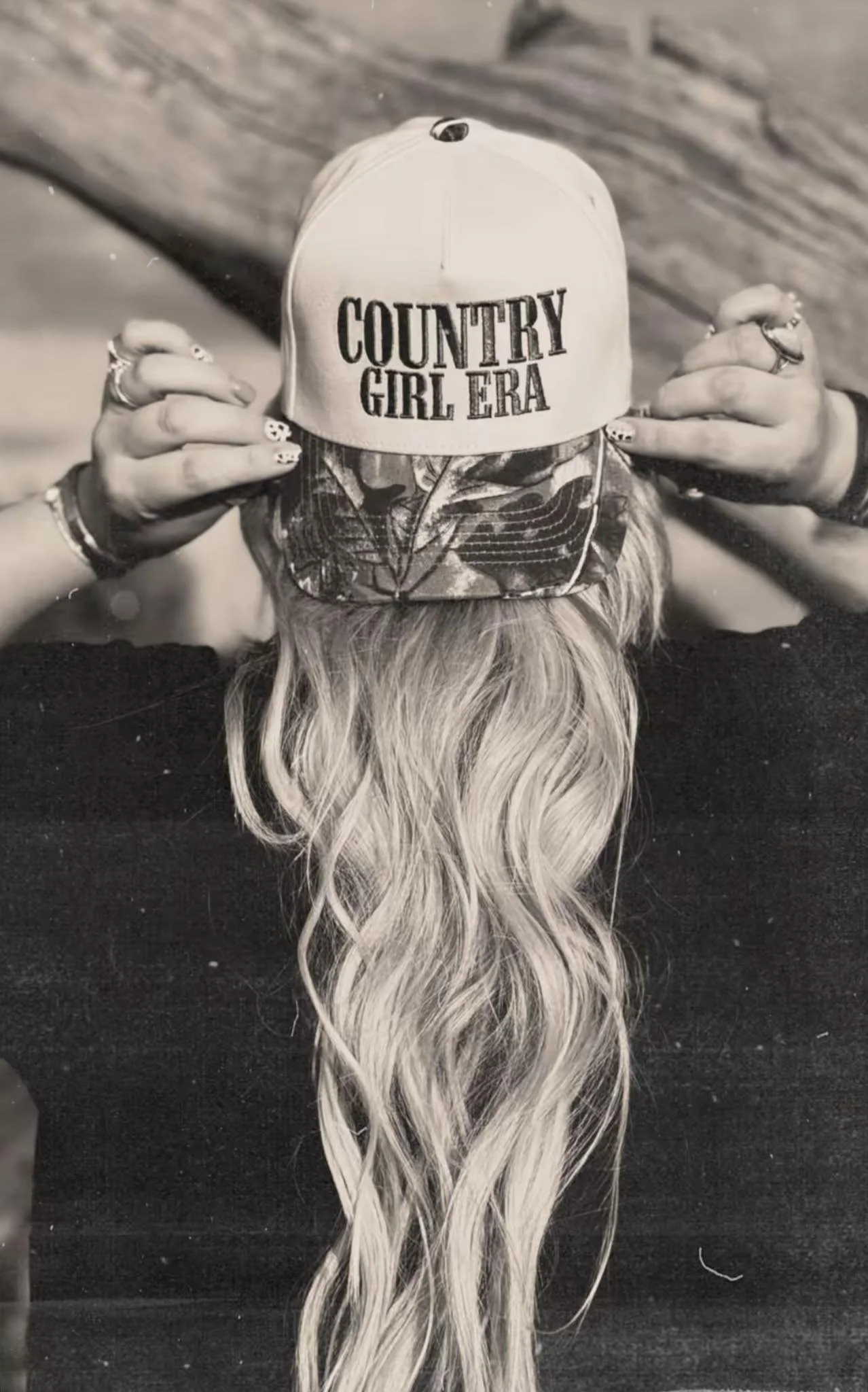 Black-and-white portrait of Gemma adjusting her country-style cap, with long blonde hair falling over her shoulders.