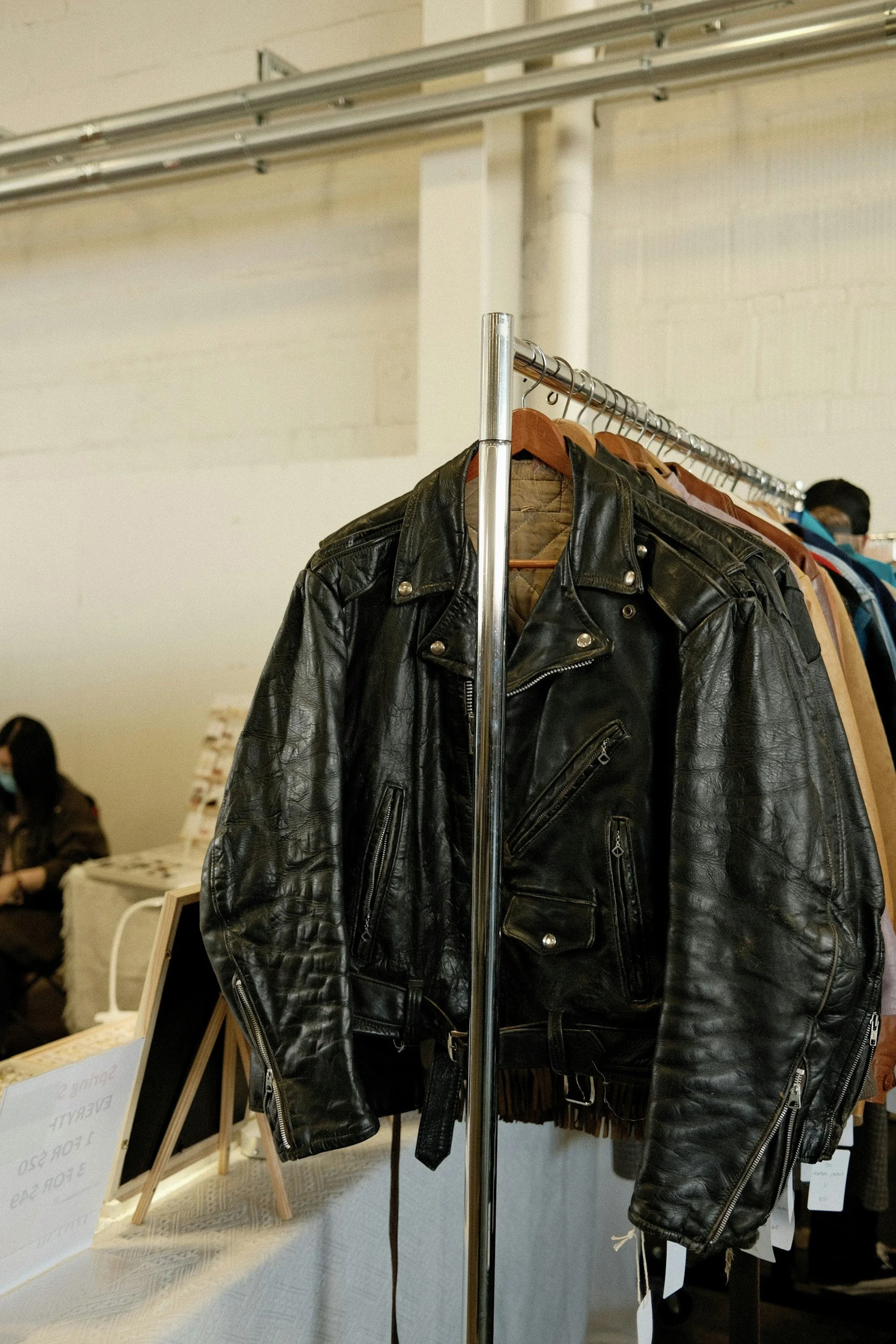 Black leather jacket hanging on a rack at a clothing market or store with signs and customers in the background.