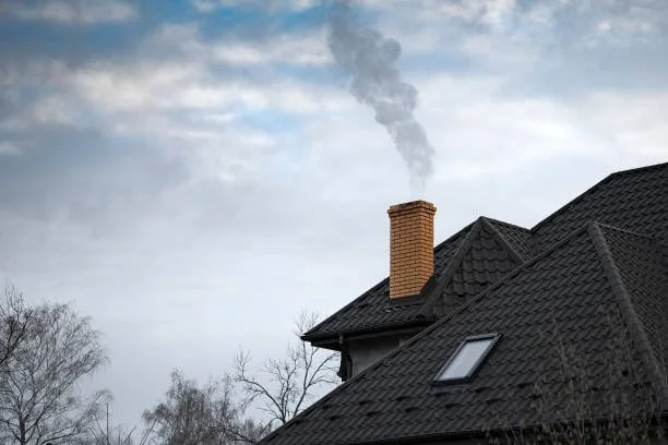 A house with a black tiled roof and a brick chimney emitting smoke, with leafless trees in the background and a cloudy sky.