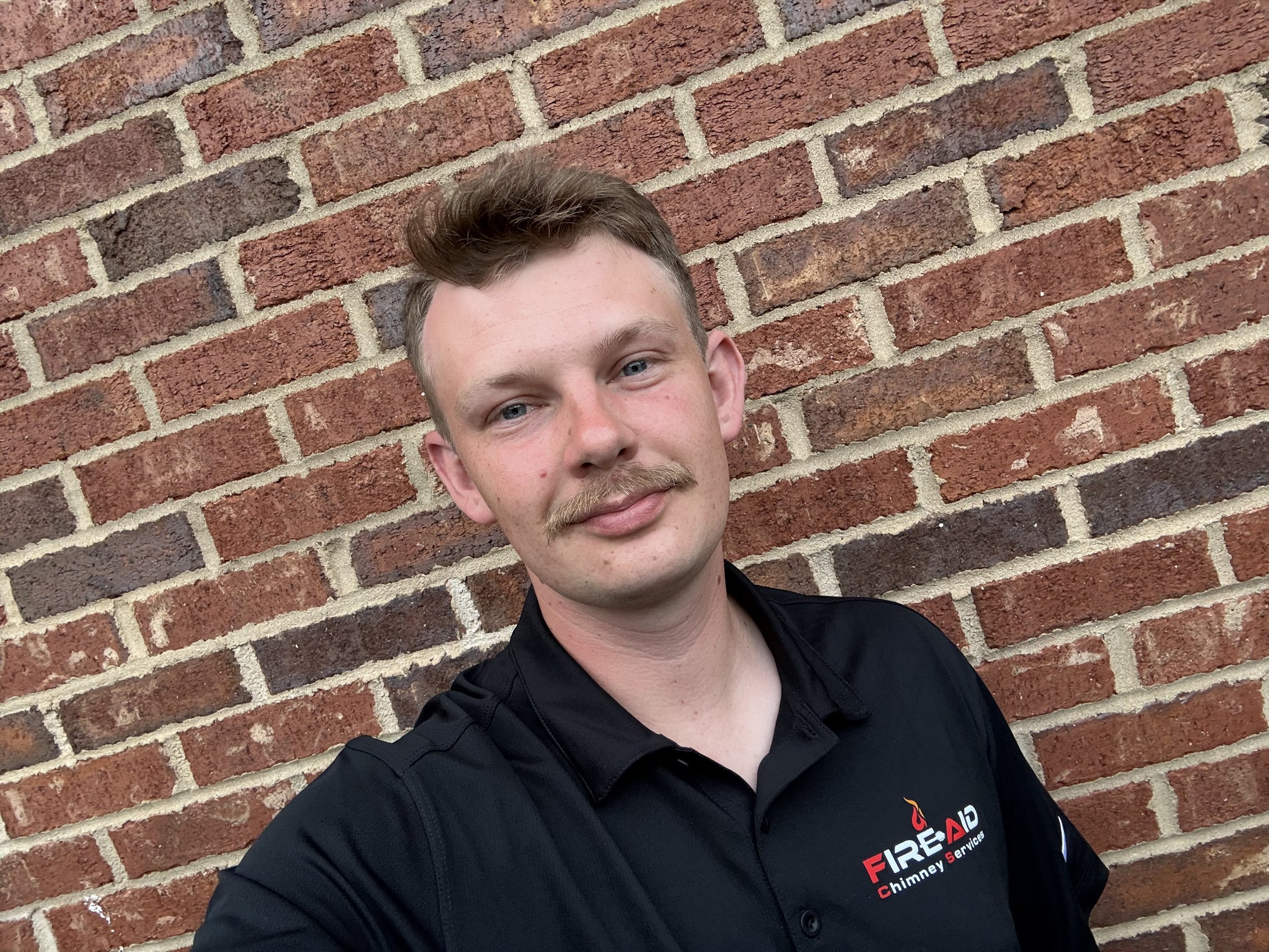 A young man with light skin, short brown hair, and facial hair smiling slightly, standing in front of a brick wall, wearing a black collared shirt with a logo on it.
