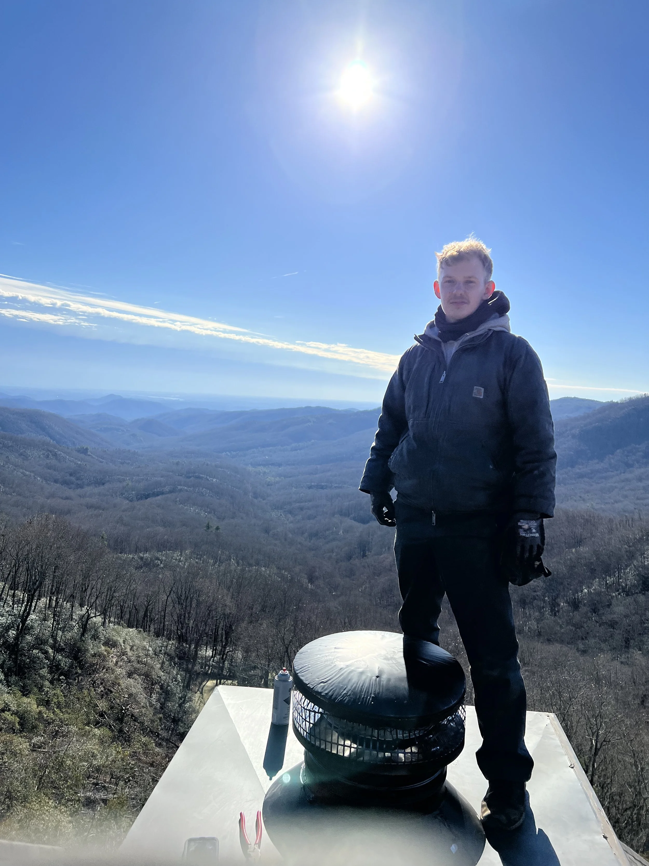 A man standing on a mountain top during daytime, wearing a dark jacket and gloves, with scenic mountain landscape in the background and the sun shining brightly in the clear blue sky.