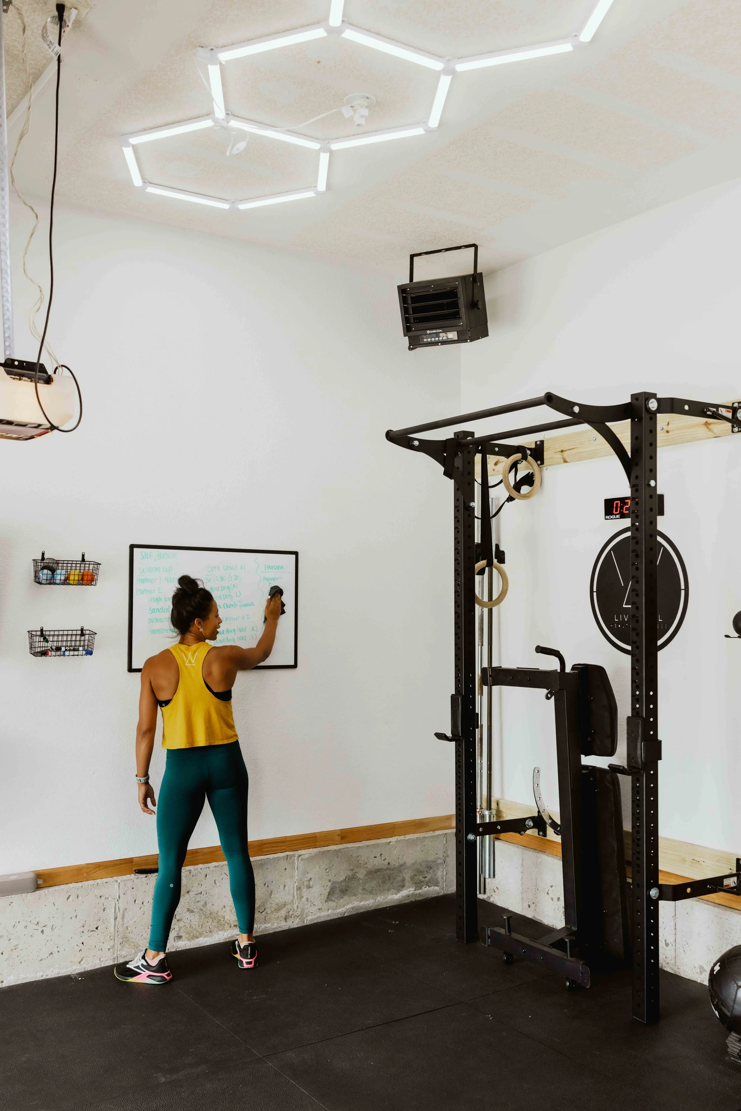A woman in athletic clothing writing on a whiteboard in a gym with fitness equipment and a hexagonal ceiling light.