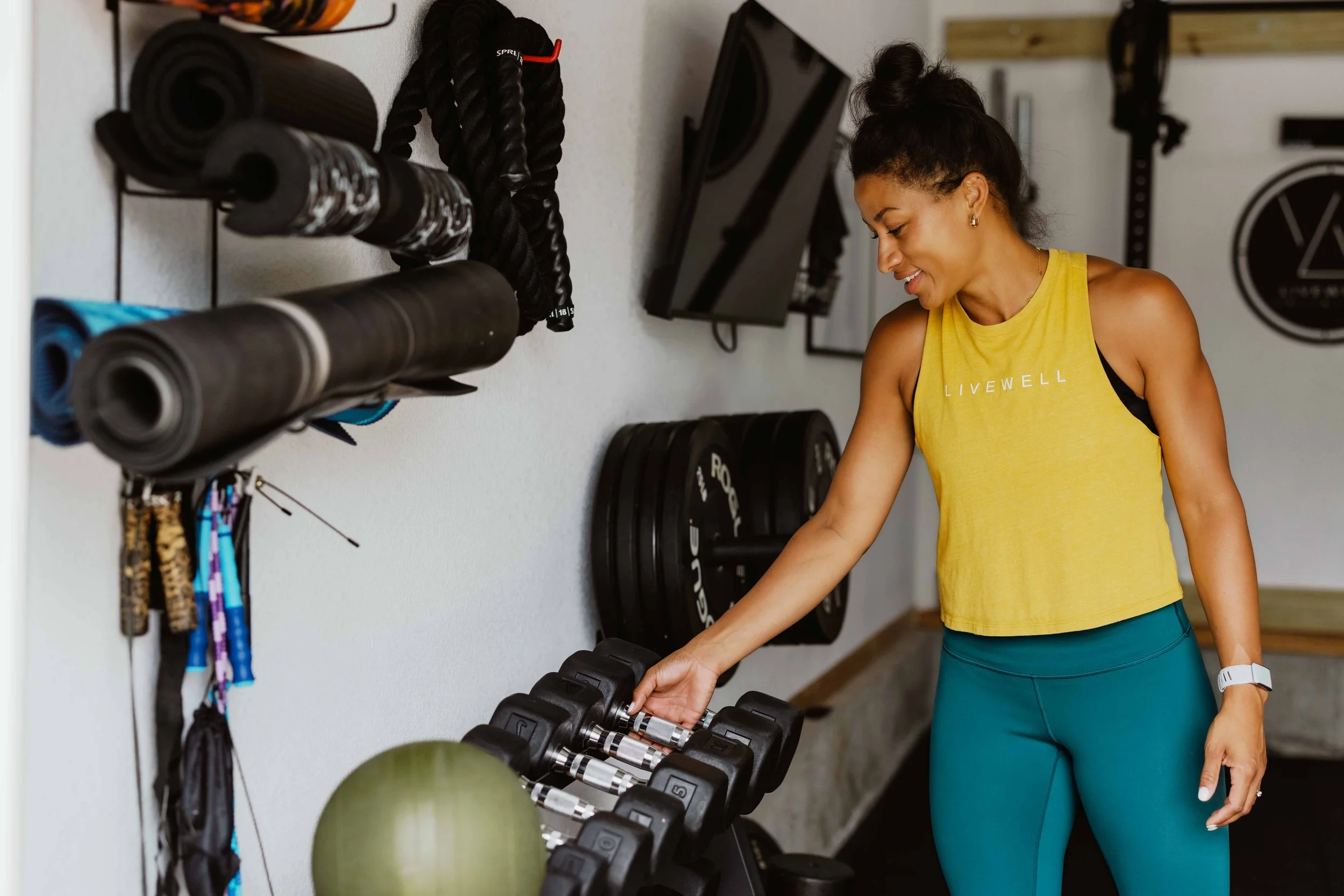 A woman with dark hair tied up in a bun, wearing a yellow sleeveless shirt and teal leggings, standing in a gym and smiling while reaching for dumbbells on a rack.