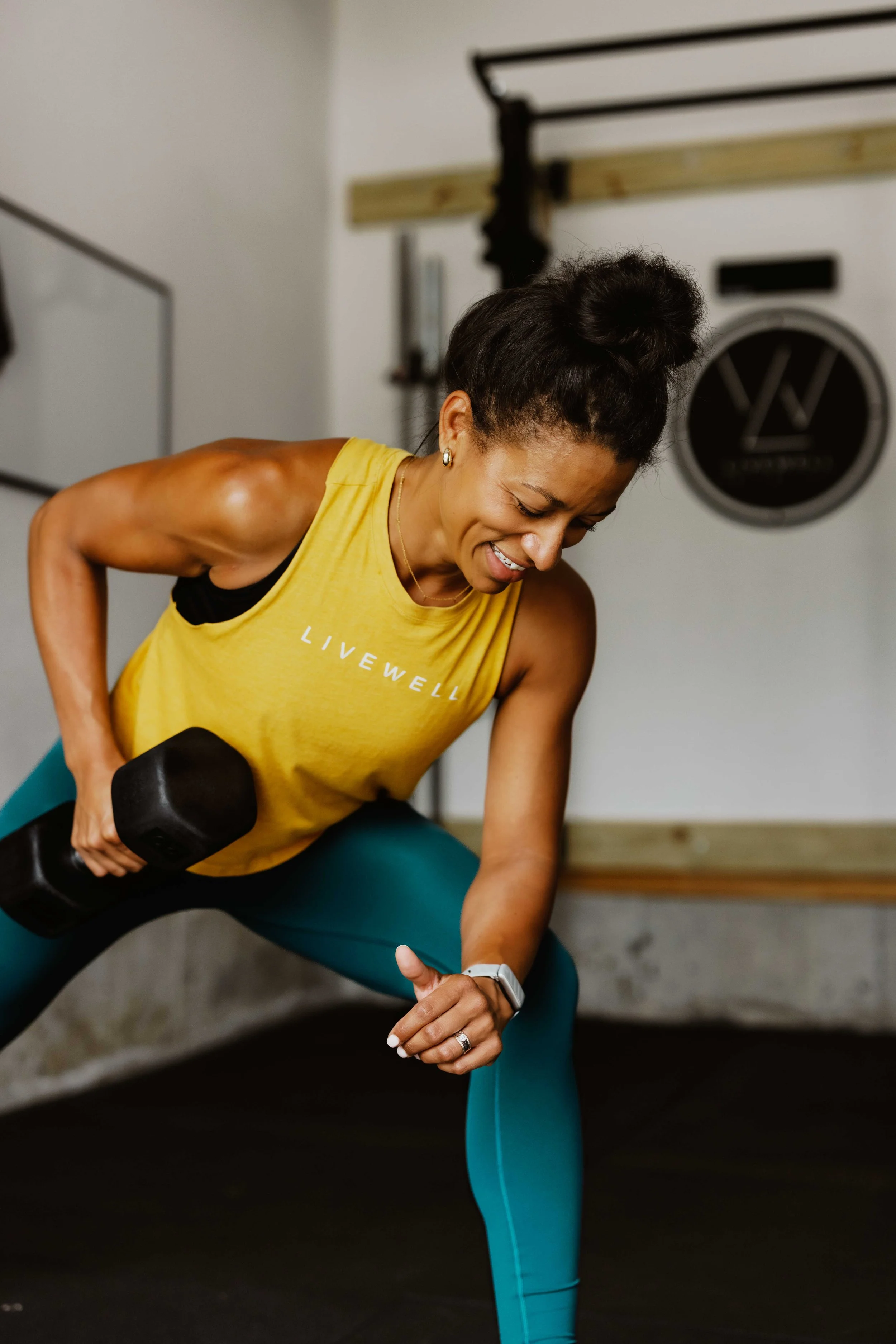 A woman with black hair tied in a bun, wearing a yellow fitness tank top and turquoise leggings, is performing a side lunge exercise while holding a black dumbbell in her right hand. She is smiling and looking down, in a gym setting with workout equipment in the background.