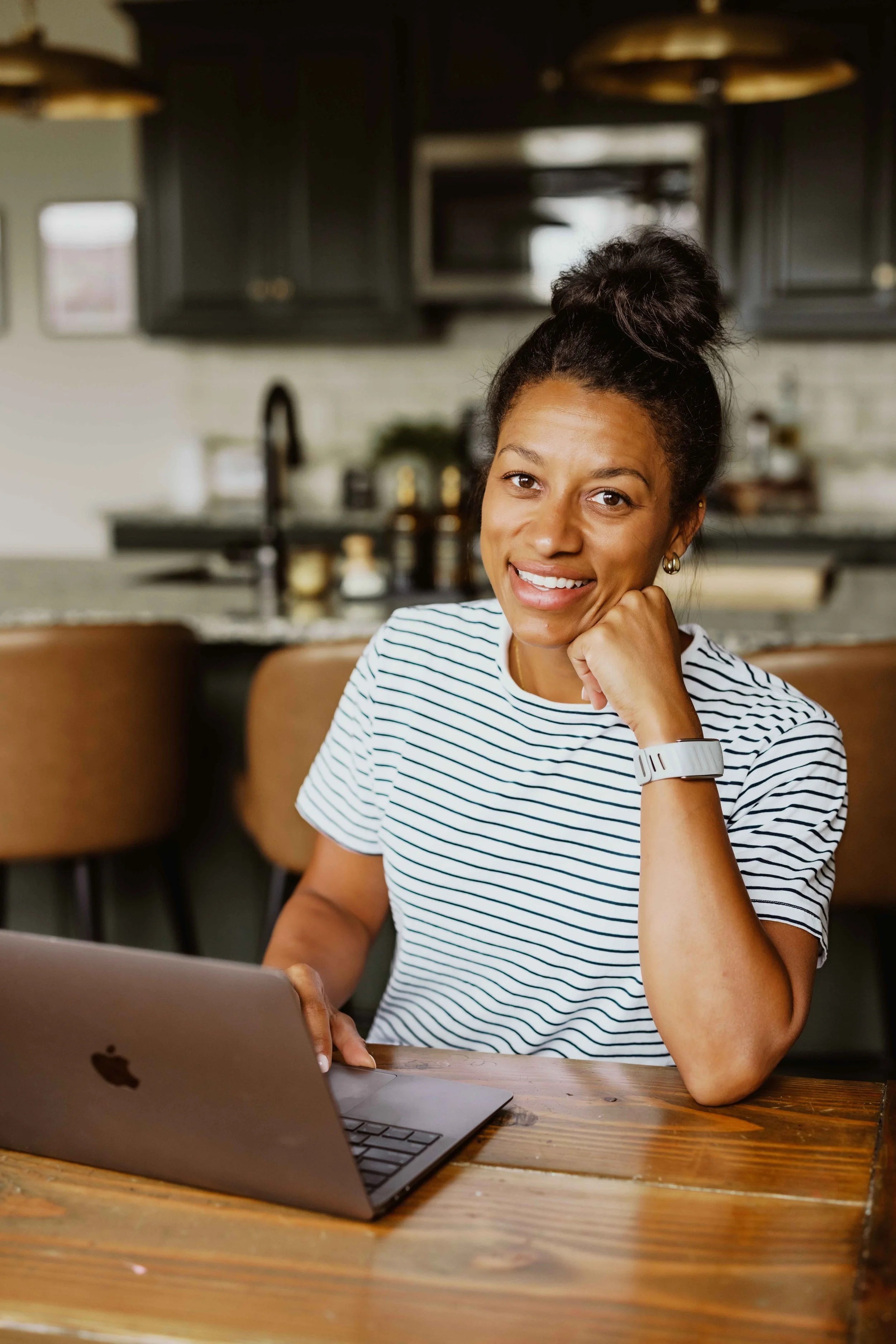 A woman with dark hair in a bun, wearing a striped shirt and earrings, sitting at a wooden table with a laptop in front of her in a modern kitchen, smiling and resting her chin on her arm.