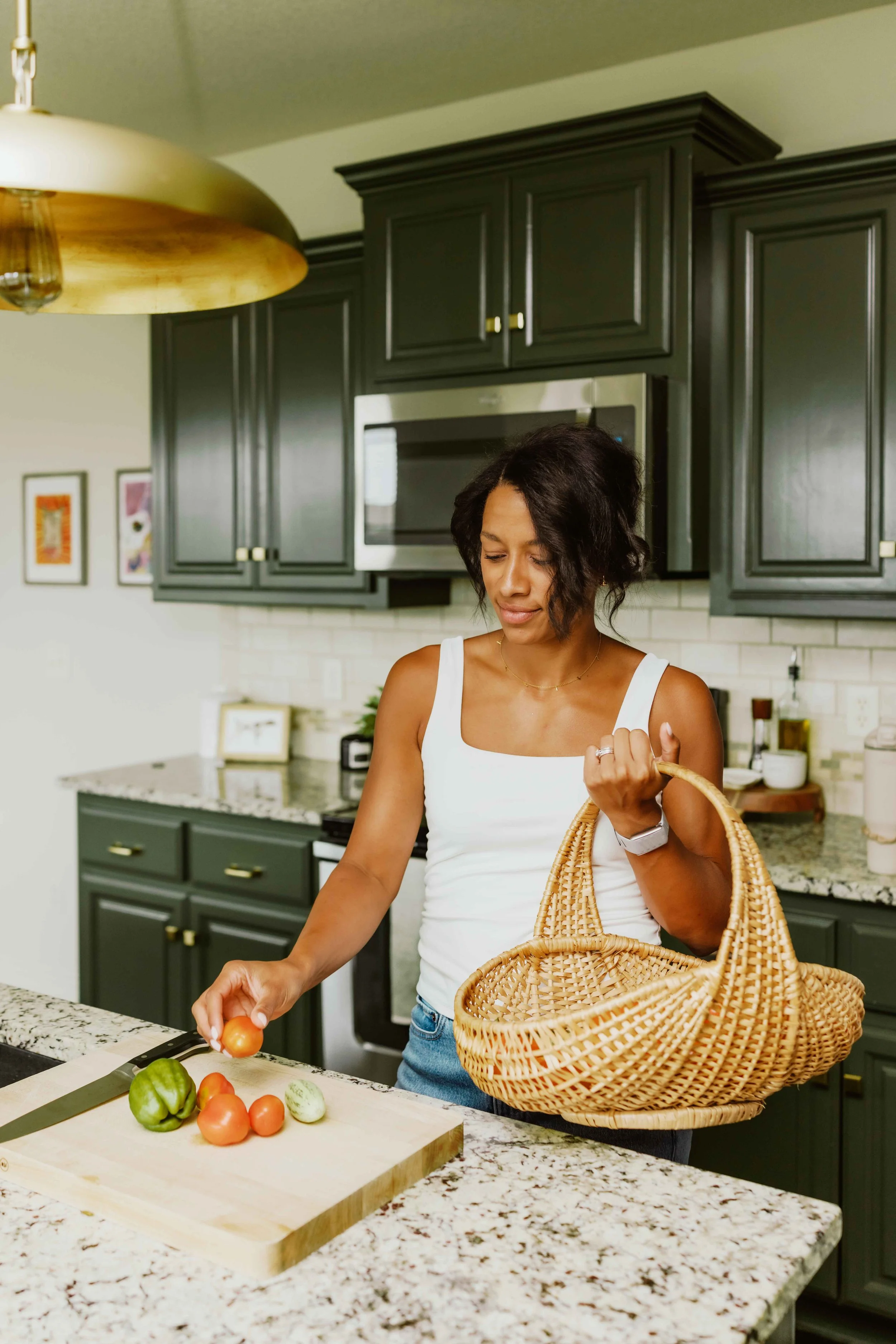 A woman in a white top prepares vegetables including tomatoes and bell peppers in a modern kitchen with black cabinets and a granite countertop. She is holding a woven basket.