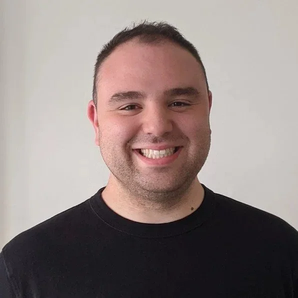 A young man with short dark hair smiling at the camera, wearing a black t-shirt, standing against a plain light-colored background.