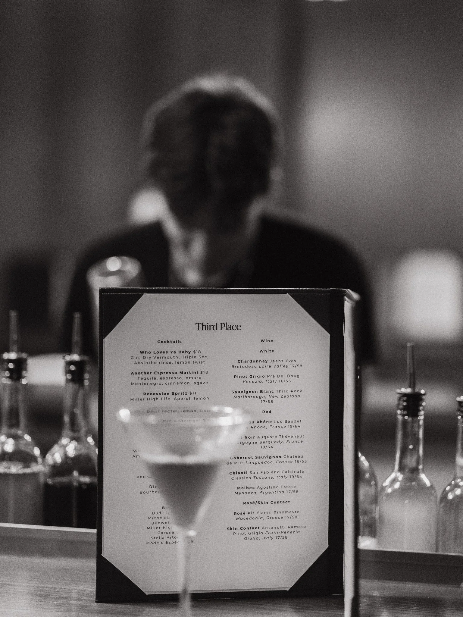A black and white photo of a table with alcohol bottles in the foreground and a menu on a stand in the background. The menu shows a third place award with details of various drink options like cocktails and wines.