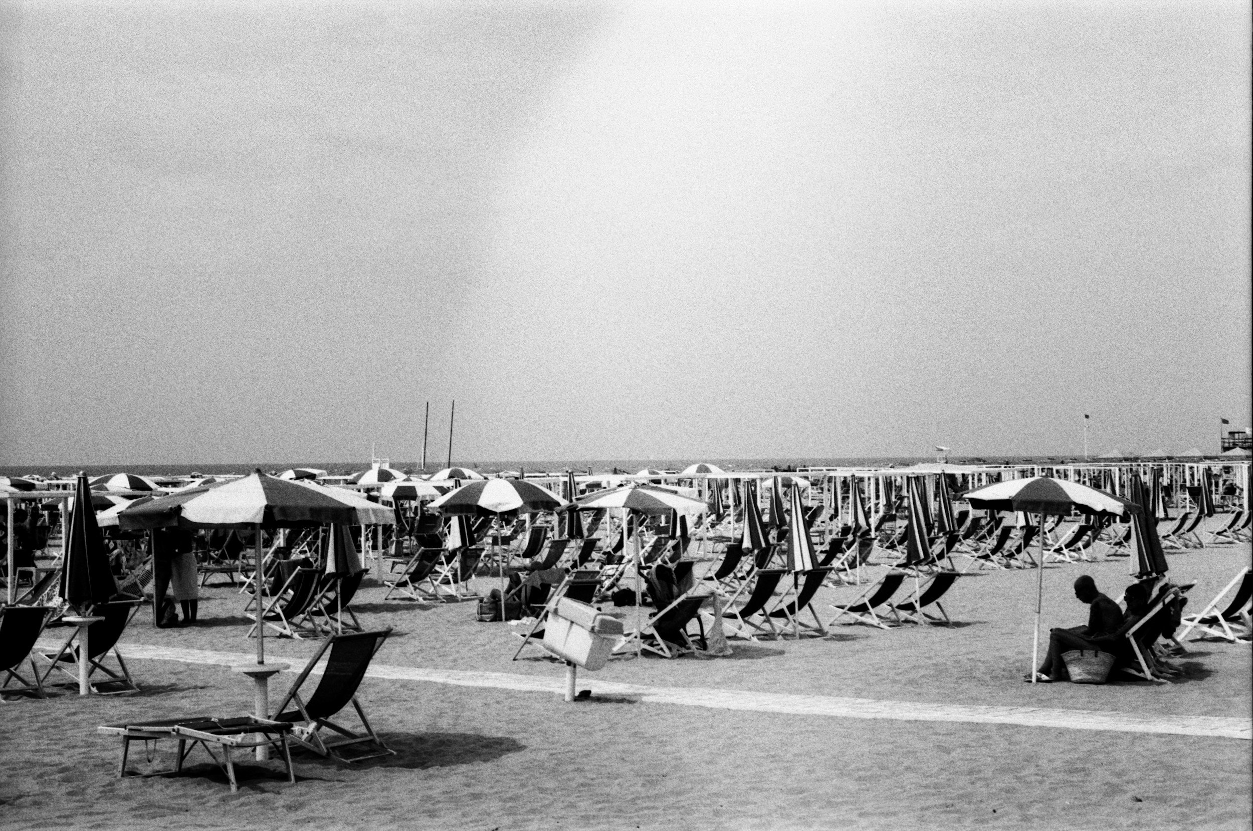 A black and white photo of a crowded beach with numerous striped umbrellas and lounge chairs. In the background, there are a few people walking, likely along the shoreline, under a clear sky.