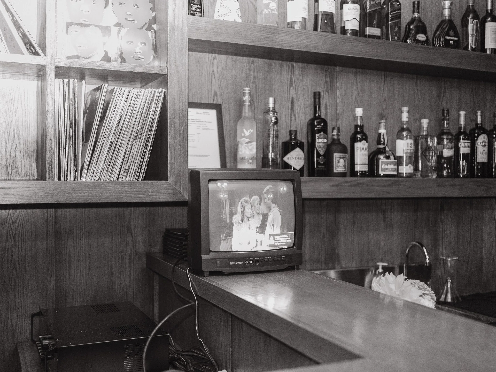 A black and white photo of a wooden shelf in a bar or restaurant, featuring liquor bottles, a framed photograph or menu, vinyl records, and a vintage television set.