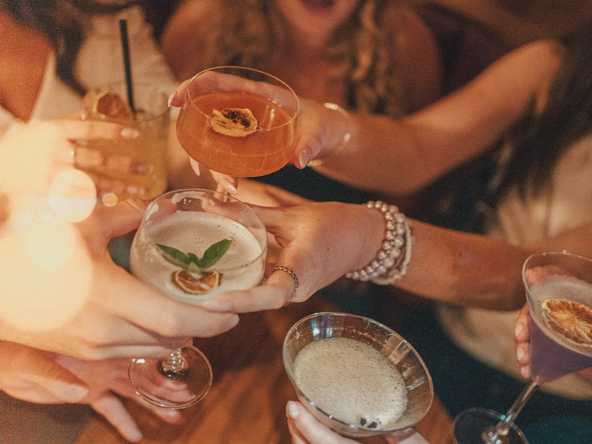 A person holding a cocktail glass with an orange-colored drink garnished with a dried fruit slice, surrounded by other drinks and party glasses, at a celebration or gathering.