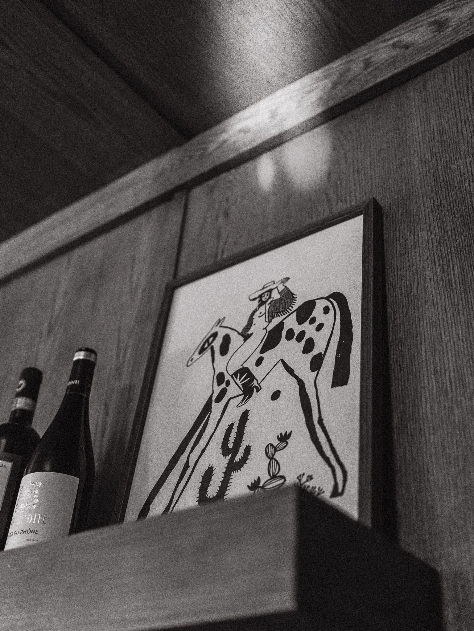 Black and white photo of a framed picture on a wooden wall, with bottles of wine or liquor beneath it.