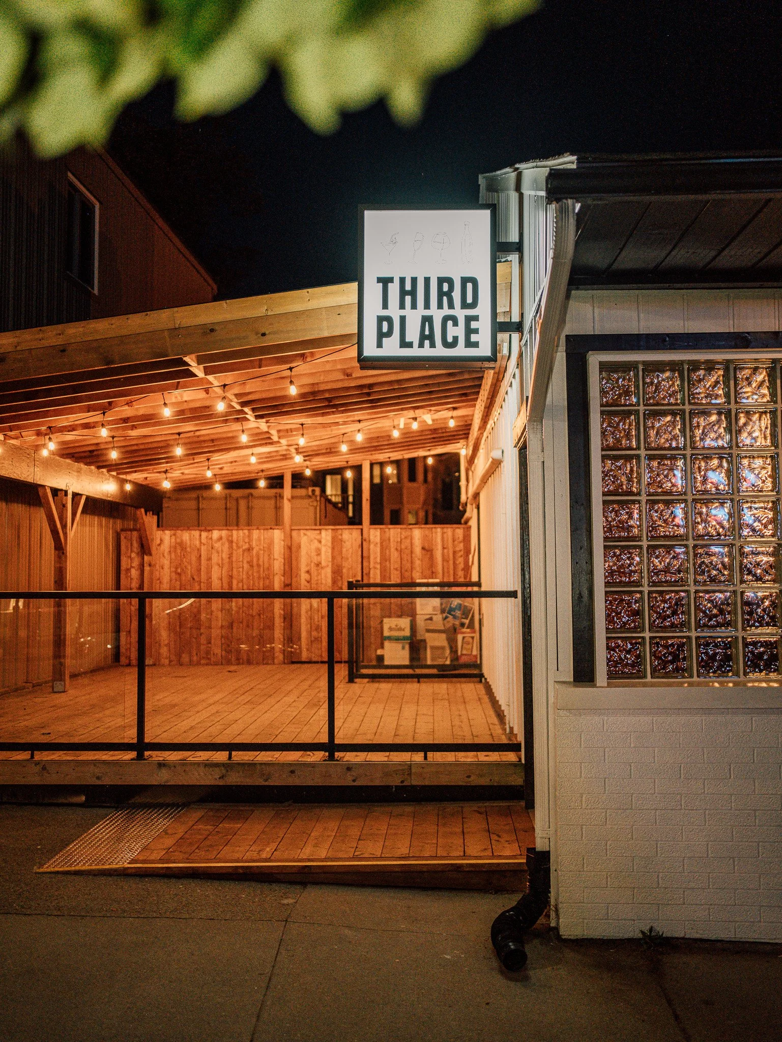 Nighttime outdoor patio area with wooden decking, string lights, and a sign that reads 'Third Place'. The patio is bordered by a wooden fence, and a building with glass block windows is visible on the right side.
