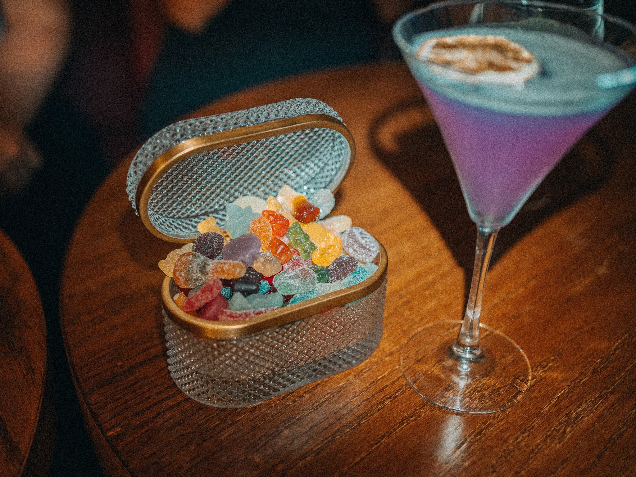 A silver jewelry box filled with colorful gummy candies, next to a martini glass with a creamy drink garnished with a cinnamon stick on a wooden table.
