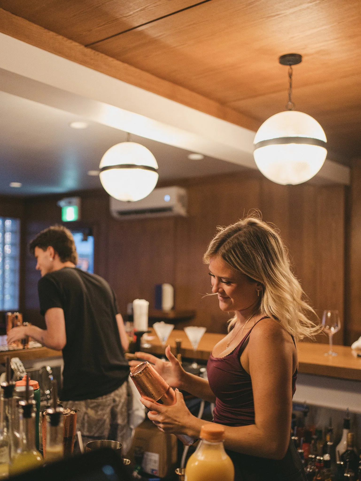 A woman at a bar holding a copper cocktail shaker, with a bartender in the background preparing drinks. The setting is a dimly lit, cozy bar with wooden accents and hanging globe lights.
