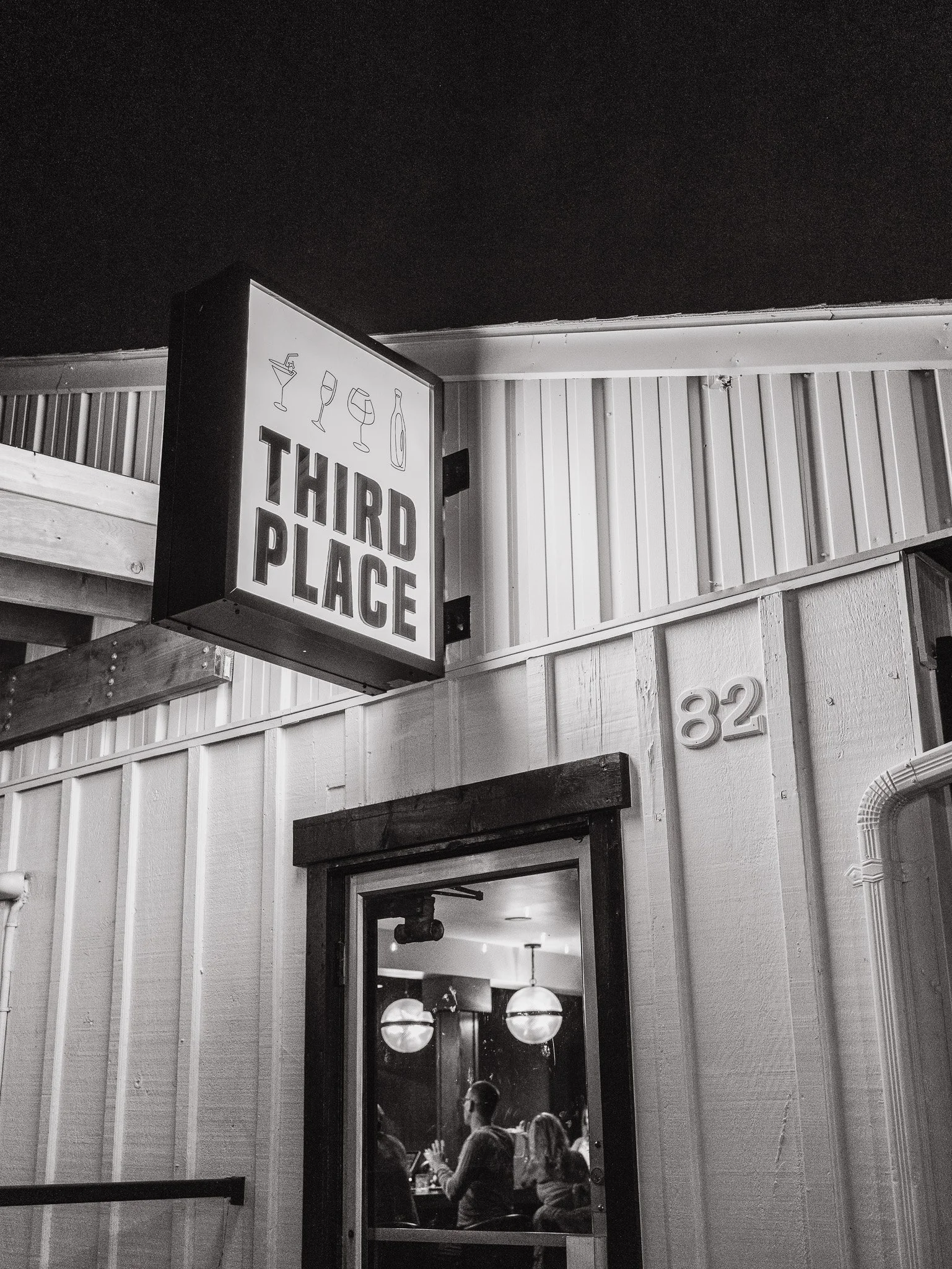 A black and white photo of the interior of a bar or restaurant with a sign that reads 'Third Place' and shows outlines of four different drinks on it. Inside, there are people, hanging globe lights, and a doorway leading into another room.