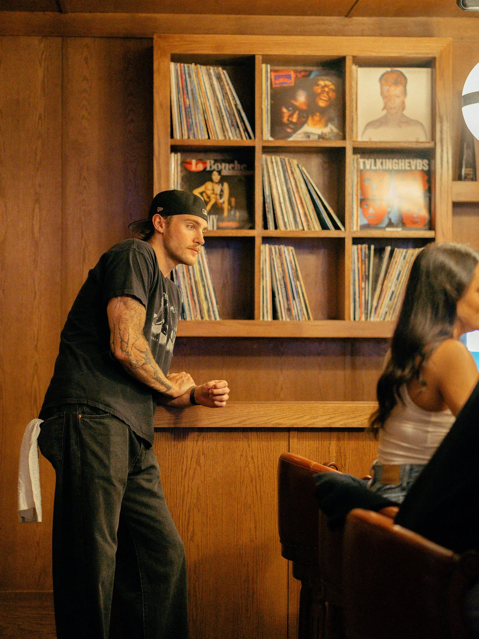 A young man with long hair, wearing a black baseball cap backward, black T-shirt, and jeans, leaning against a wooden bar or counter, with a wooden bookshelf behind him filled with vinyl records and album cover posters, in a cozy indoor setting.