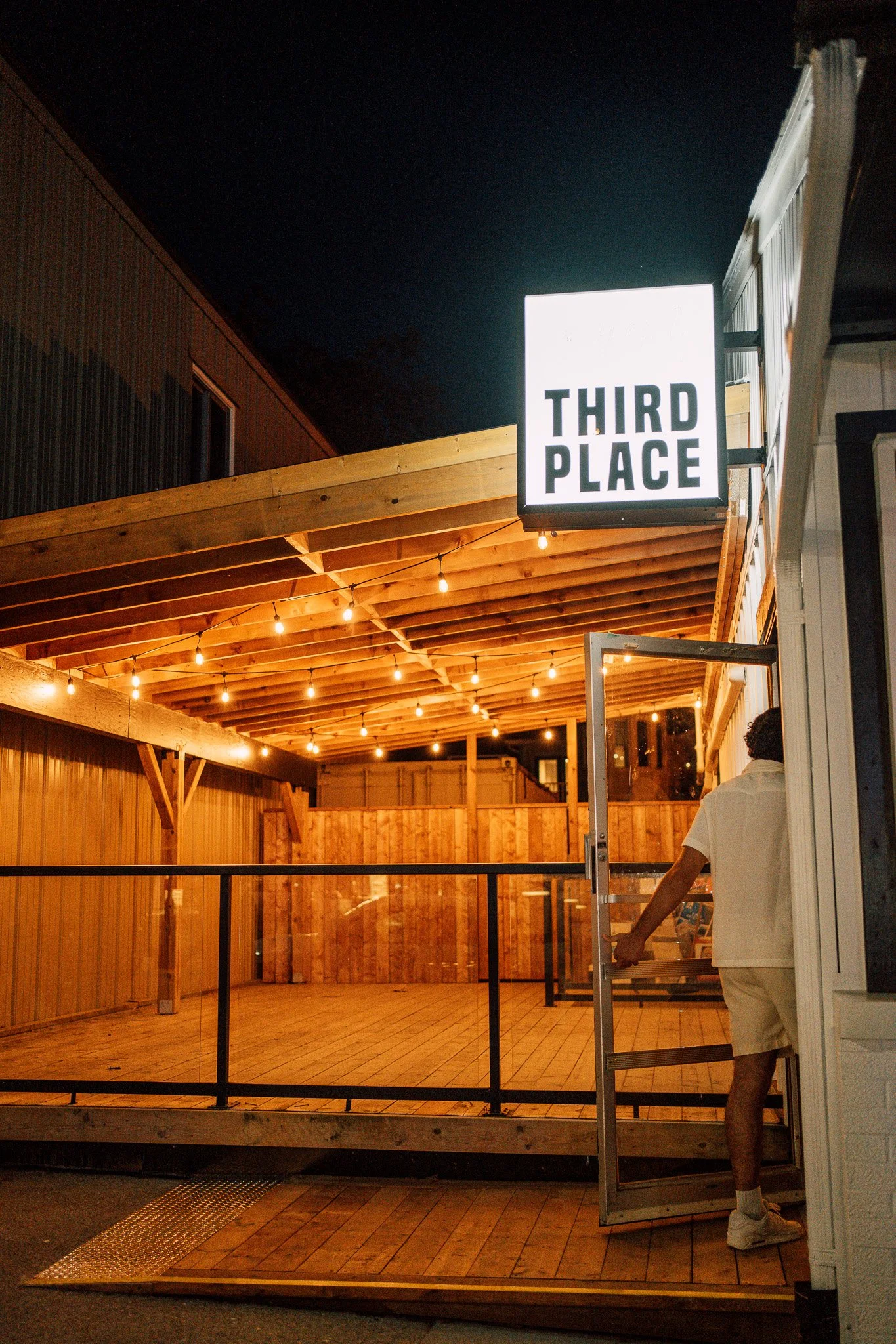 Night shot of the exterior of a building with a large illuminated sign that reads 'Third Place'. The building has wooden accents and string lights underneath the awning. A person is partially visible entering or exiting through a door.