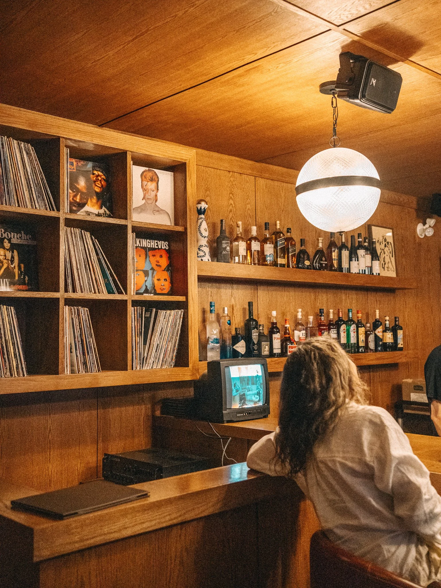 A person with curly hair wearing a white shirt seated at a wooden bar with shelves of liquor bottles and vinyl records on the wall behind. A small monitor on the counter displays a photo, and a ceiling light illuminates the scene.