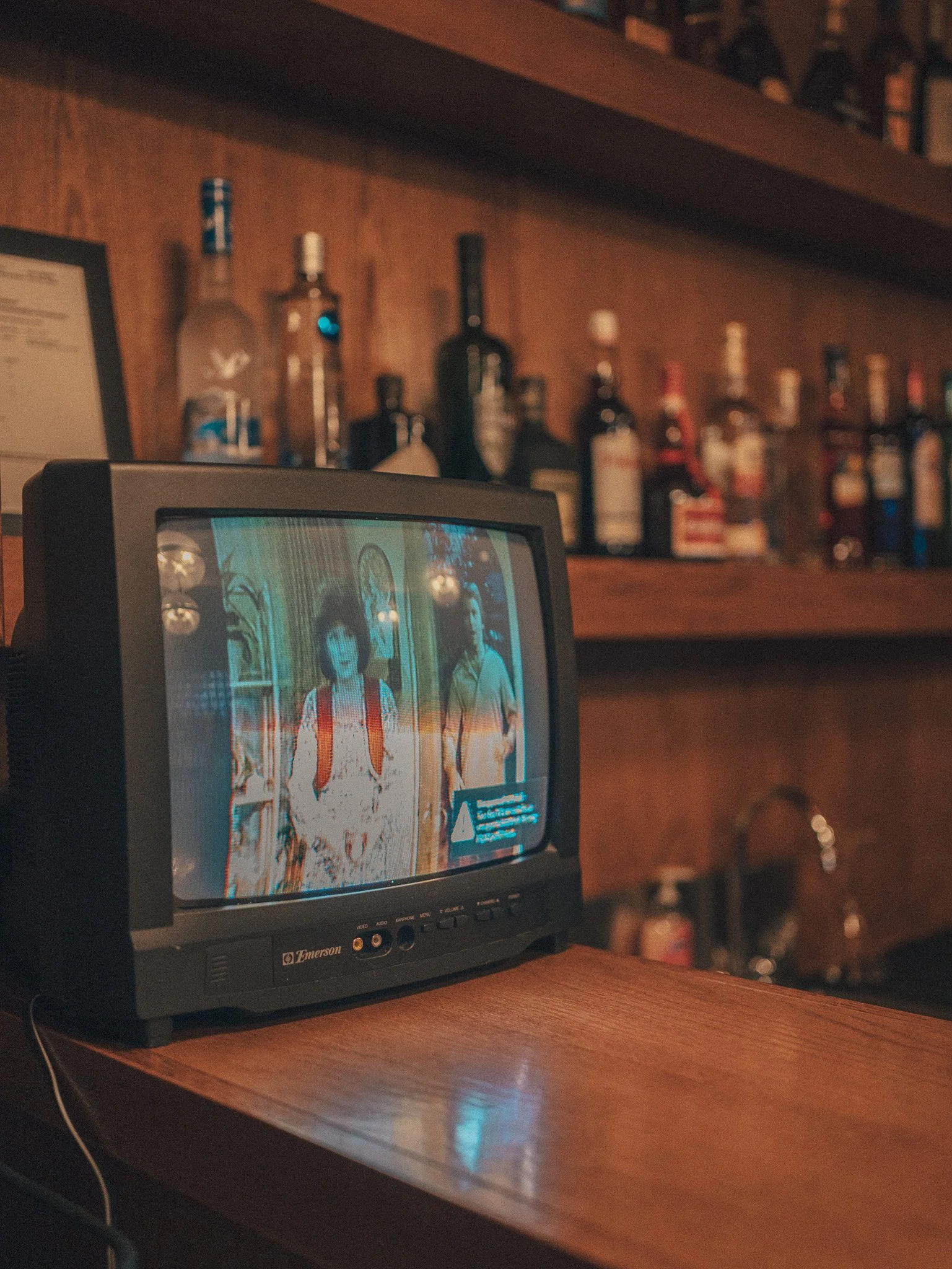 A vintage CRT TV displaying a scene from a TV show or movie of two children, one girl with dark hair and a red dress, and one boy with light hair and a light shirt, on a wooden shelf with liquor bottles in the background.